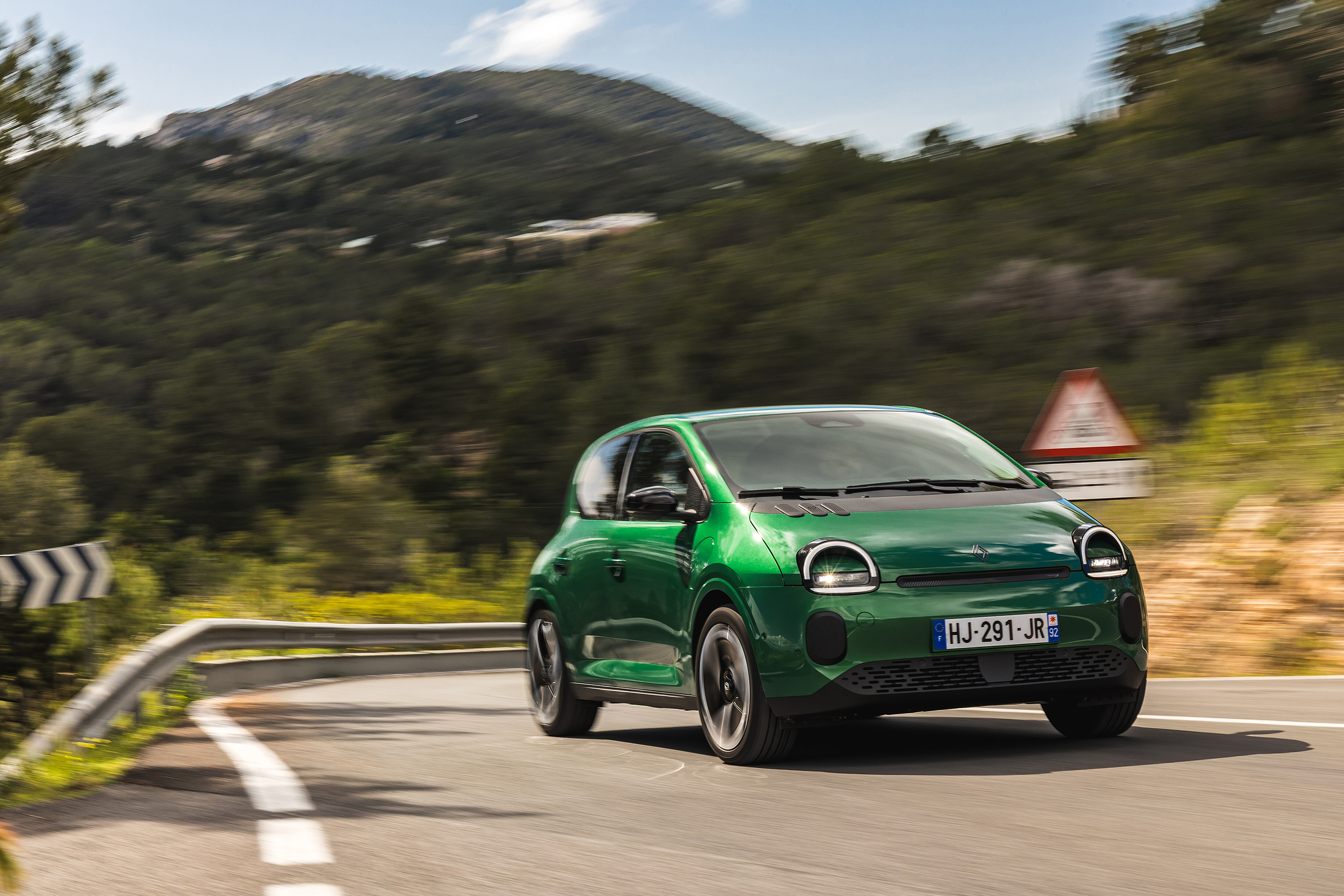 Grüner Renault Twingo mit runden Scheinwerfern fährt durch eine Kurve einer bewaldeten Bergstraße.