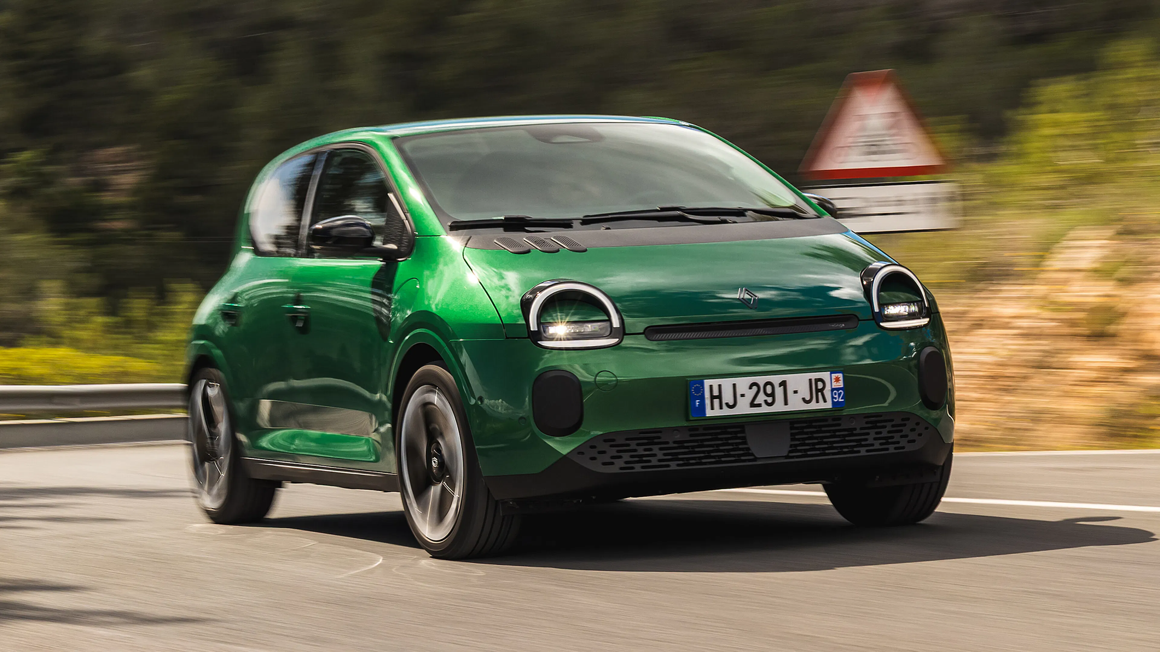 Grüner Renault Twingo mit runden Scheinwerfern fährt durch eine Kurve einer bewaldeten Bergstraße.