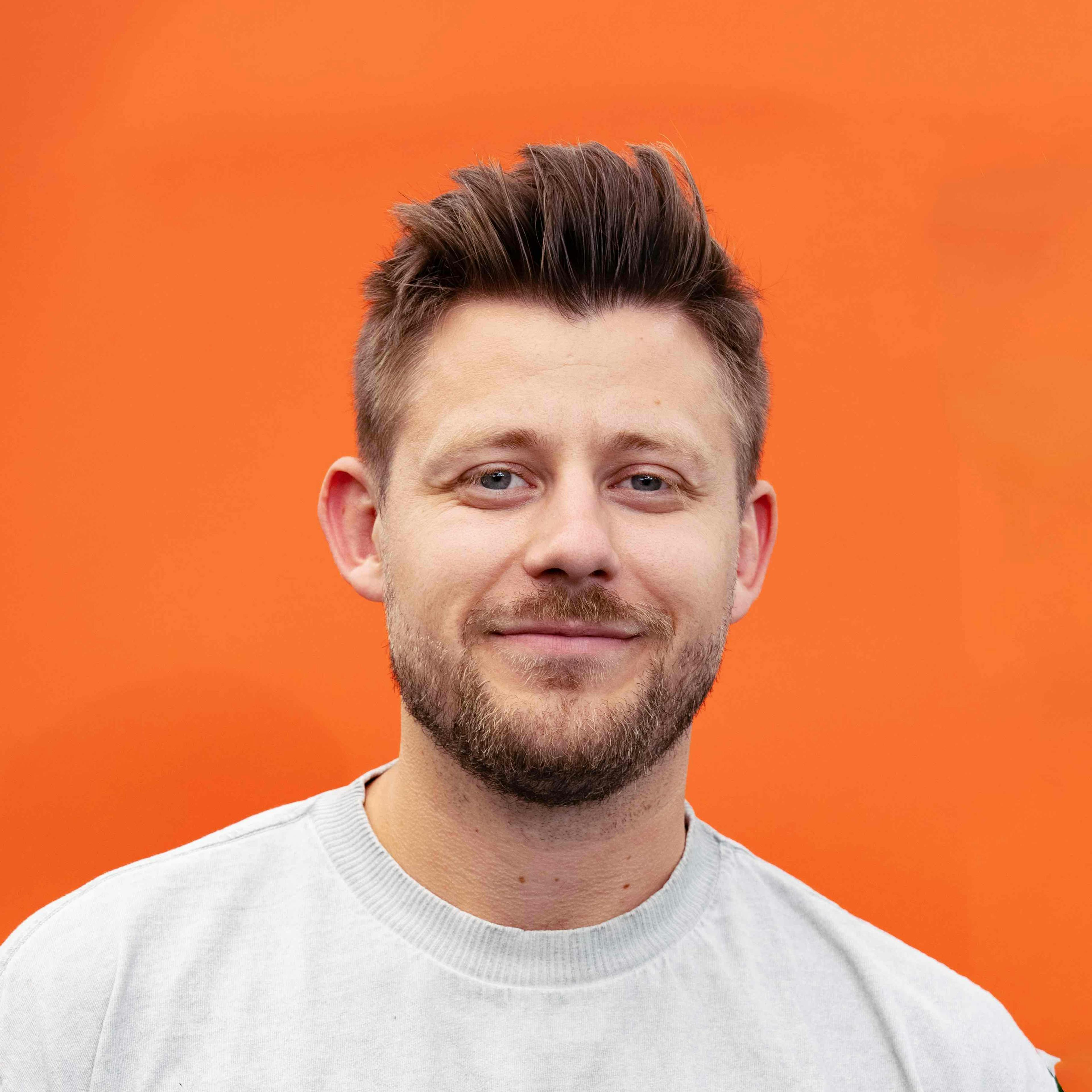 Ein lächelnder Mann mit Bart und kurz gestylten Haaren vor einem leuchtend orangen Hintergrund, trägt ein hellgraues Sweatshirt.