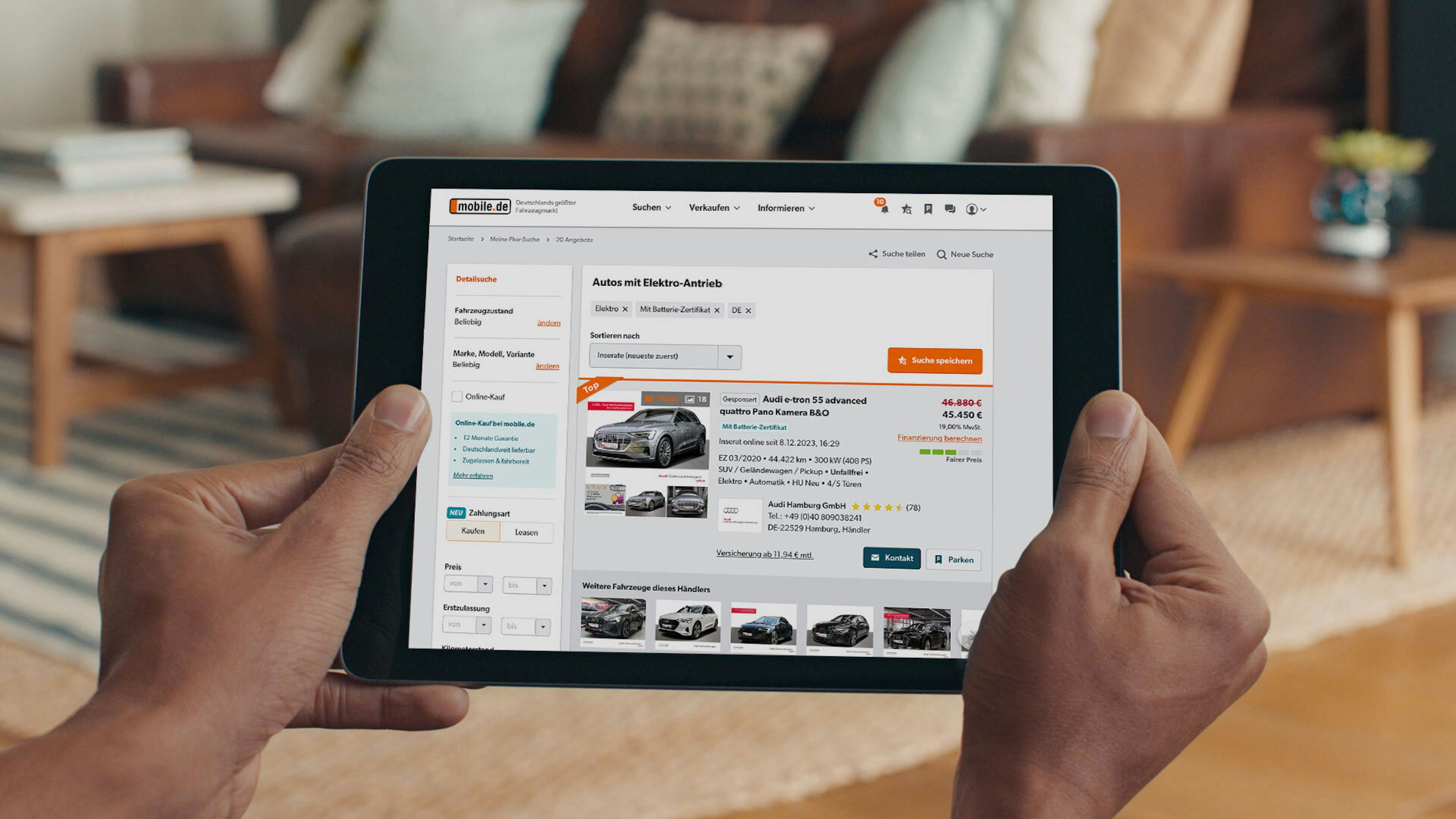 Person hält ein Tablet und durchsucht eine Autokauf-Website nach Gebrauchtwagen. Im Hintergrund ein Sofa und ein gemütliches Wohnzimmer.
