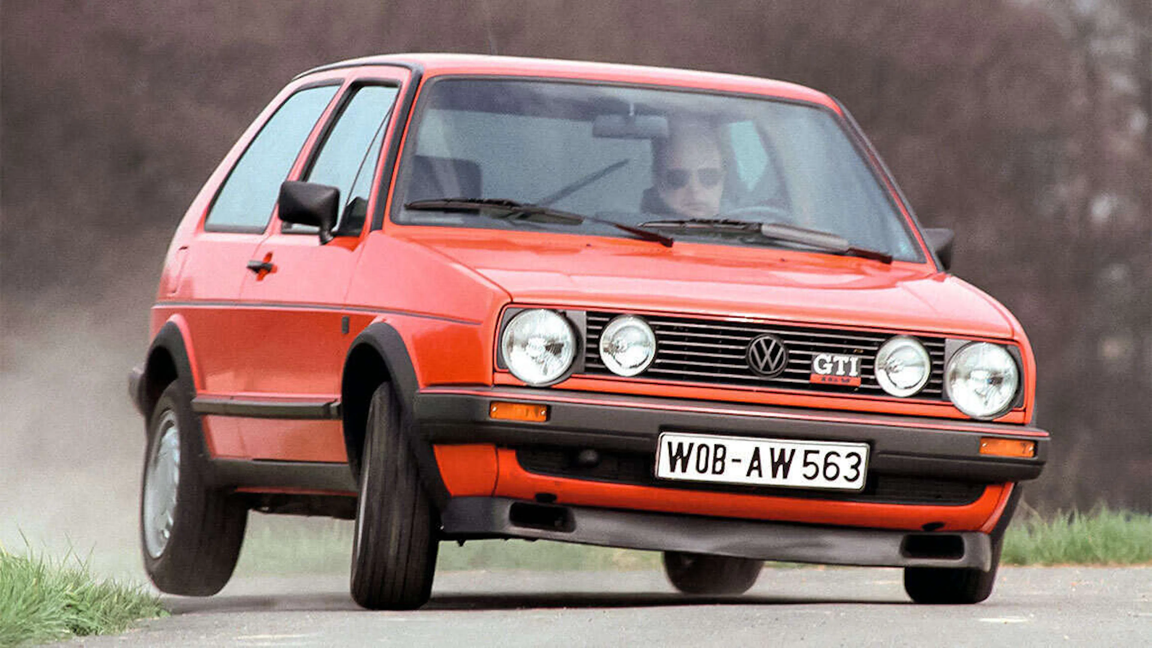 Roter VW Golf II GTI fährt dynamisch auf einer Schotterstraße, Frontansicht leicht von vorn mit Bewegungsunschärfe.