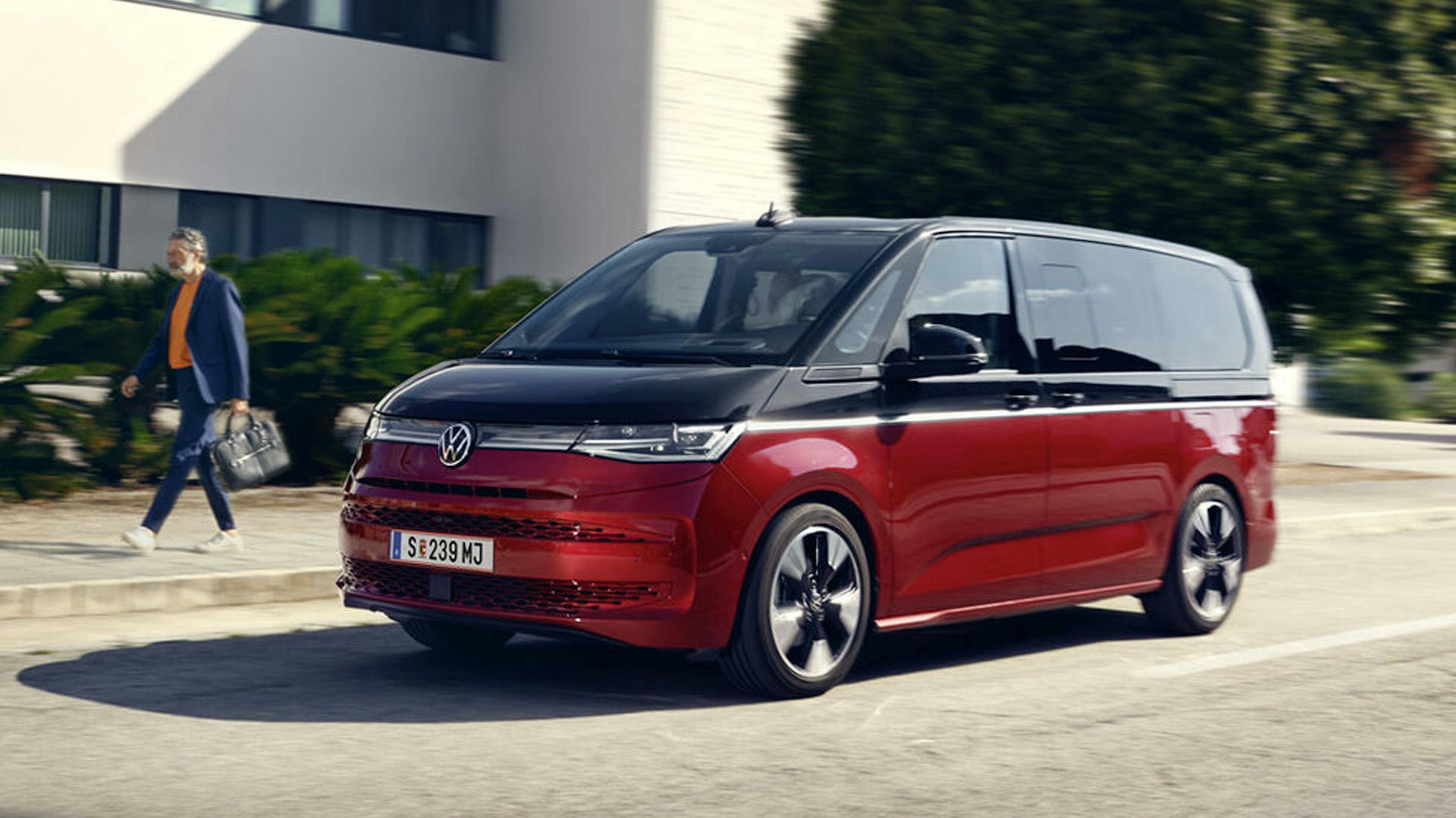 Ein schwarz-roter VW T7 eHybrid fährt an einem weißen Gebäude vorbei.