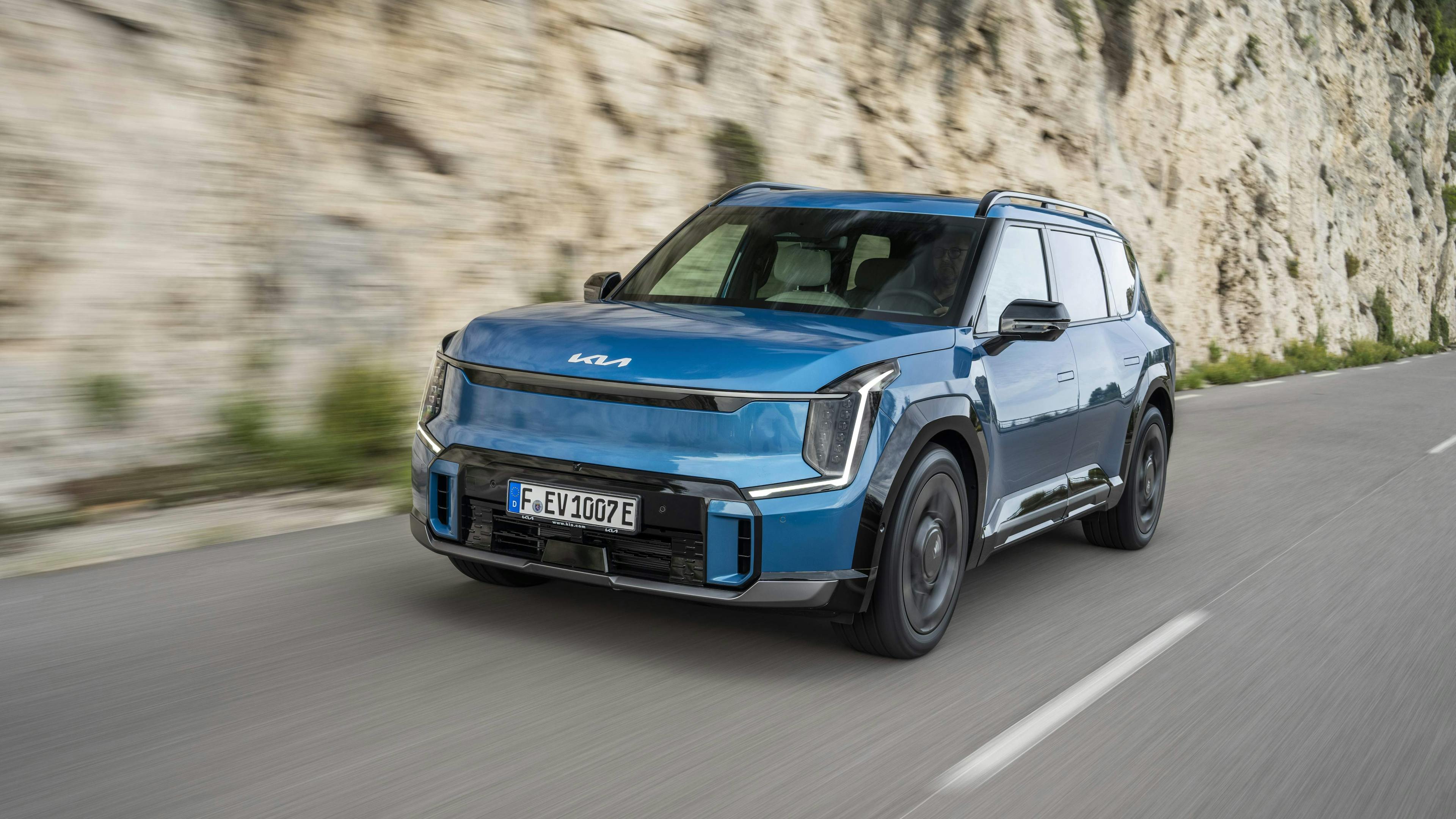 A blue Kia EV9 drives in front of a rock face on a country road.