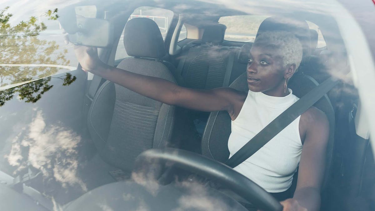 Eine Frau sitzt angeschnallt auf dem Fahrersitz eines Pkw und stellt Innenspiegel und Autositz ein.