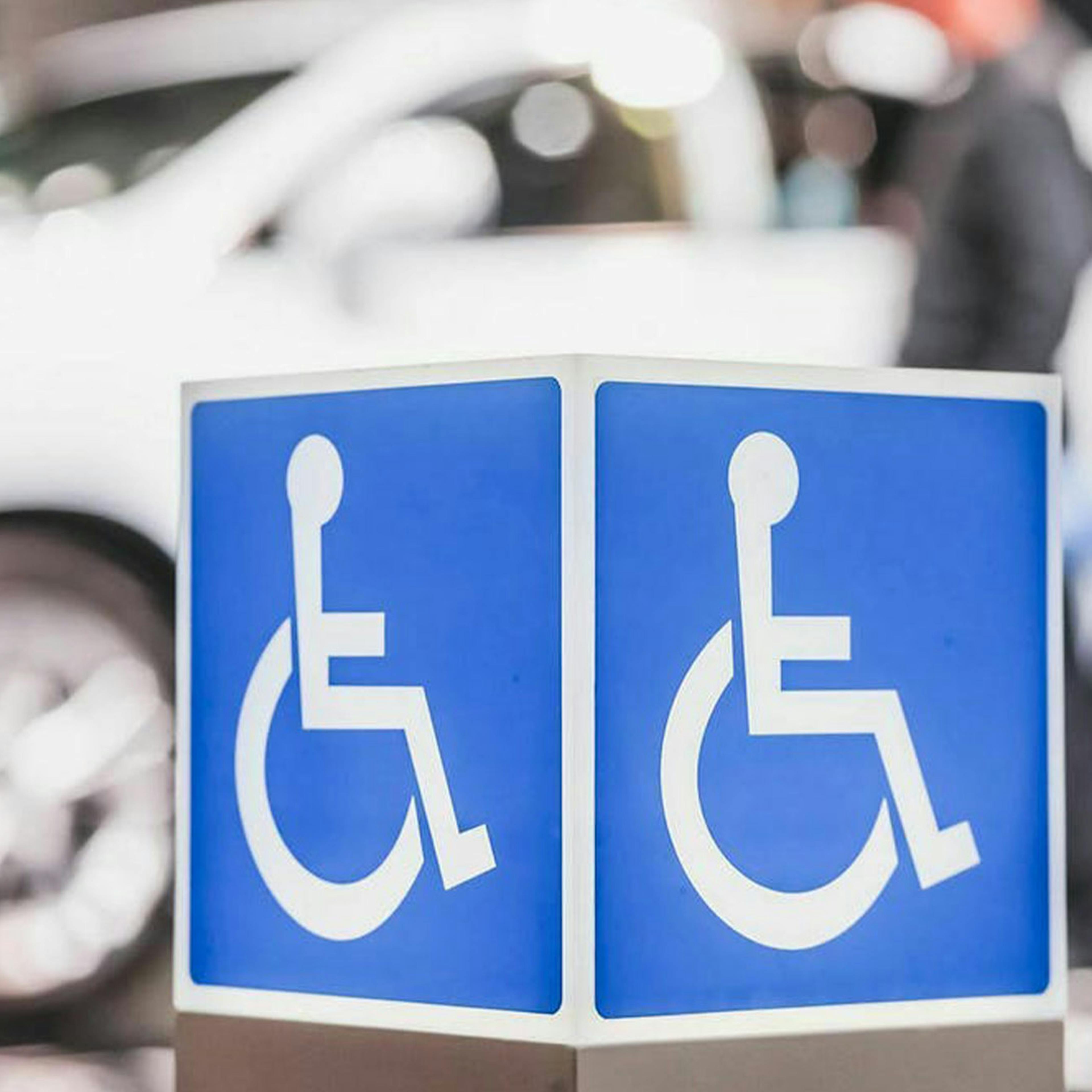 Ein weißes Auto steht hinter einem blauen Schild, auf dem ein Piktogramm mit einem Rollstuhlfahrer zu sehen ist