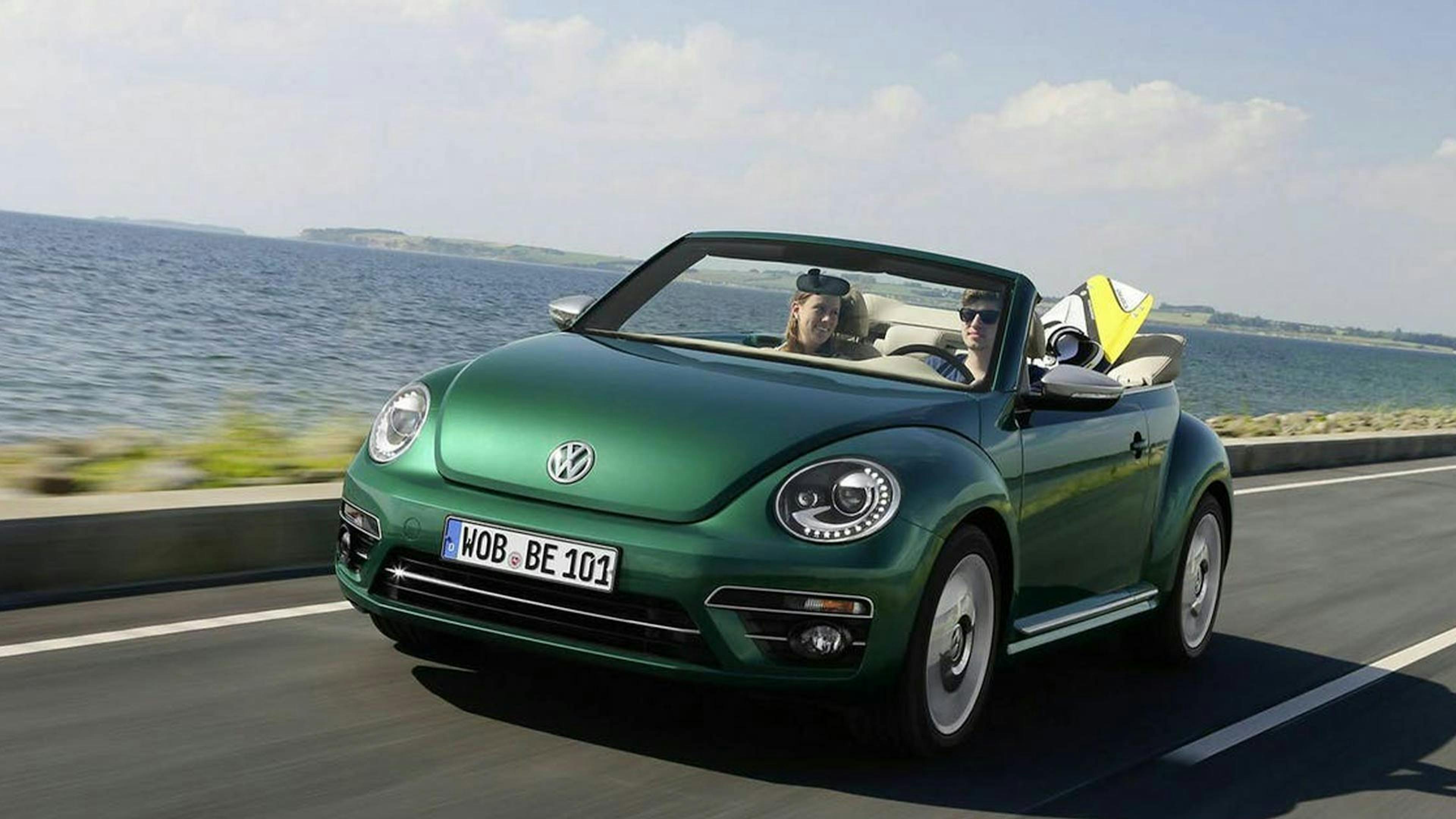 Ein grüner VW New Beetle mit geöffnetem Cabrioverdeck fährt eine Küstenstraße entlang.