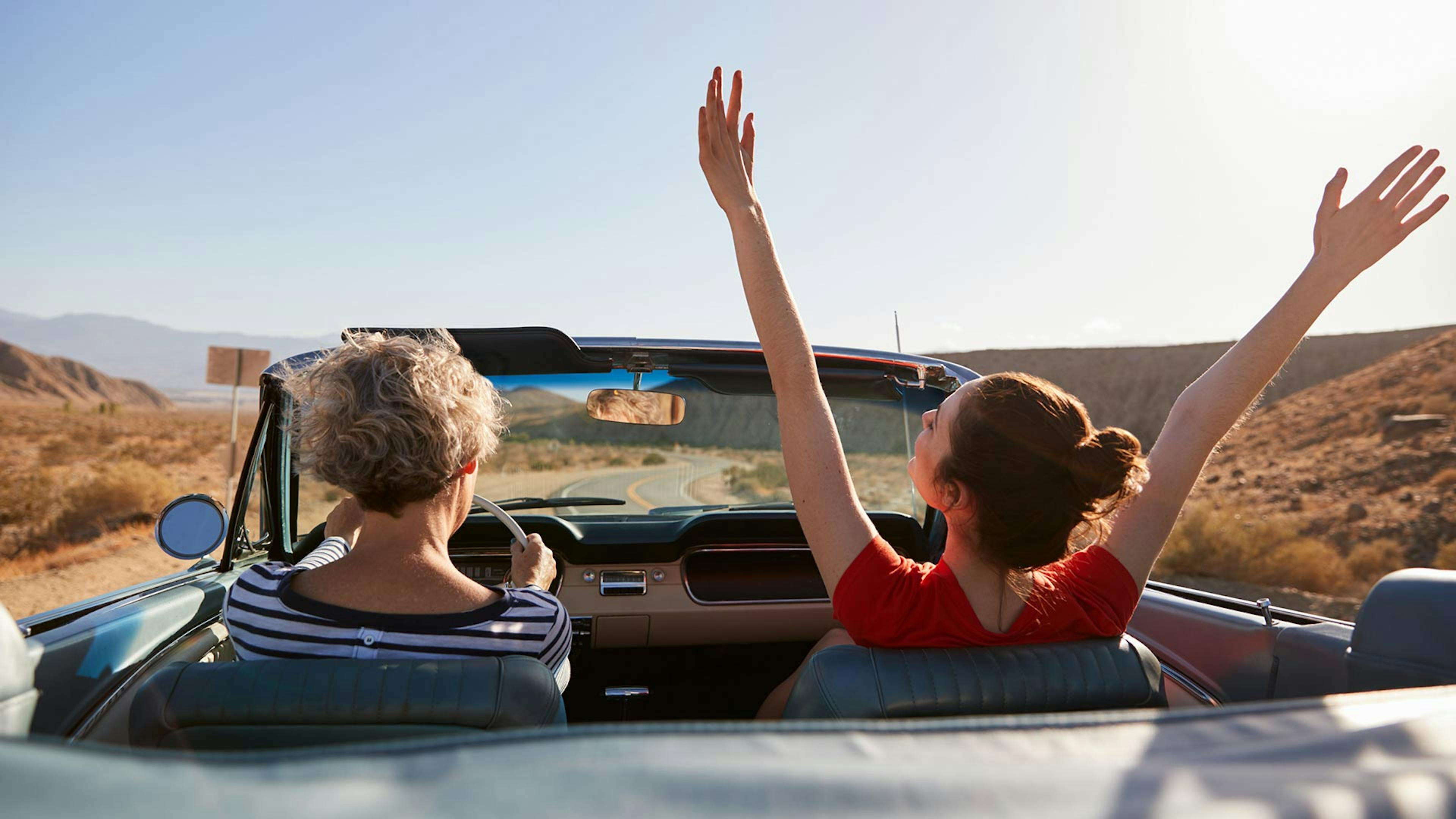 Rückansicht von zwei Frauen, die in einem Cabrio mit offenem Verdeck eine Straße entlang fahren.