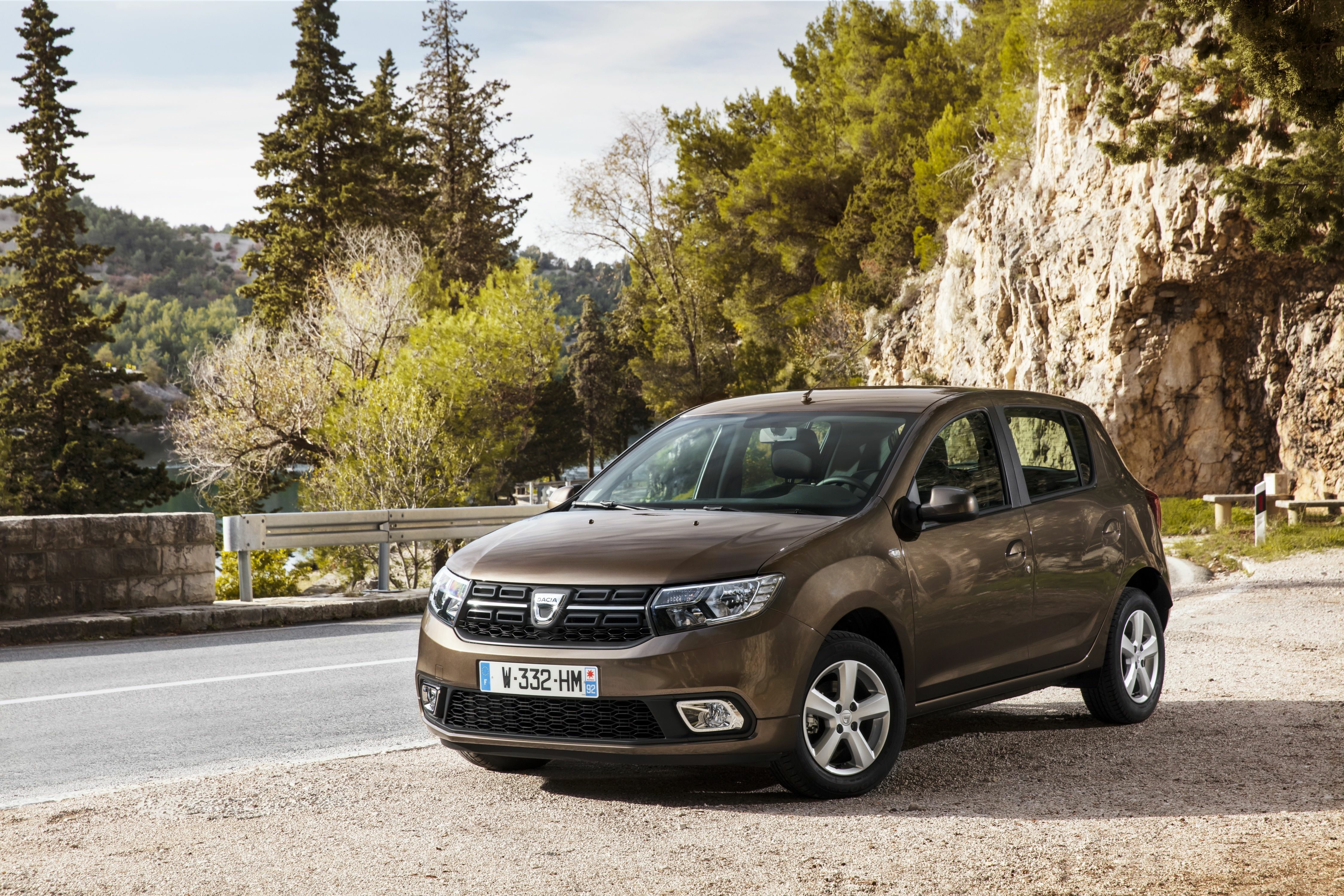 Brauner Dacia Sandero parkt in der Schrägansicht von vorn an einer Straße vor einer Felswand und Nadelbäumen.