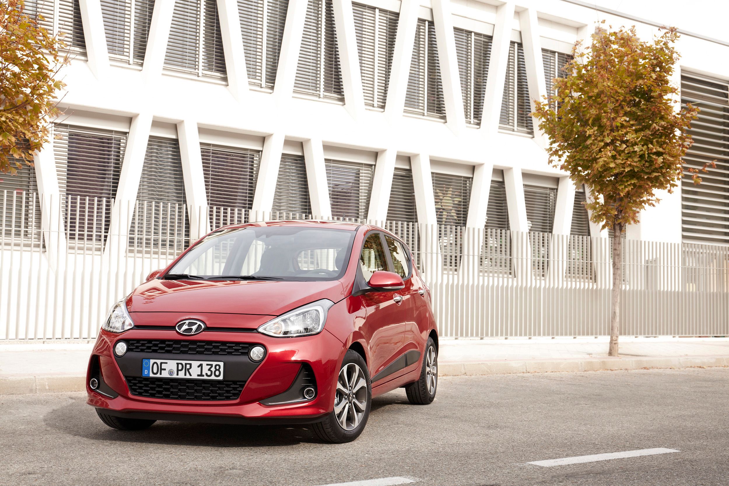 Roter Hyundai i10 steht schräg von vorn gesehen auf einer Straße vor einem weißen Gebäude mit Jalousien.