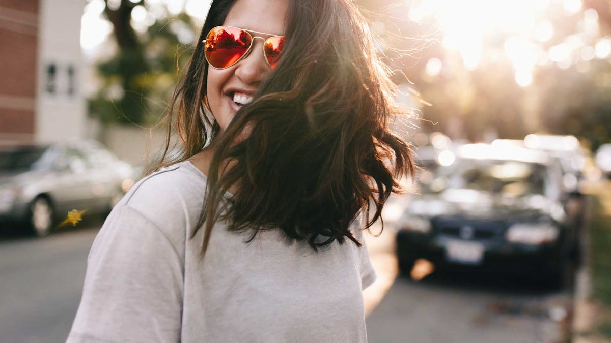 Eine junge Frau mit braunen Haaren und Sonnenbrille steht vor einem schwarzen Pkw