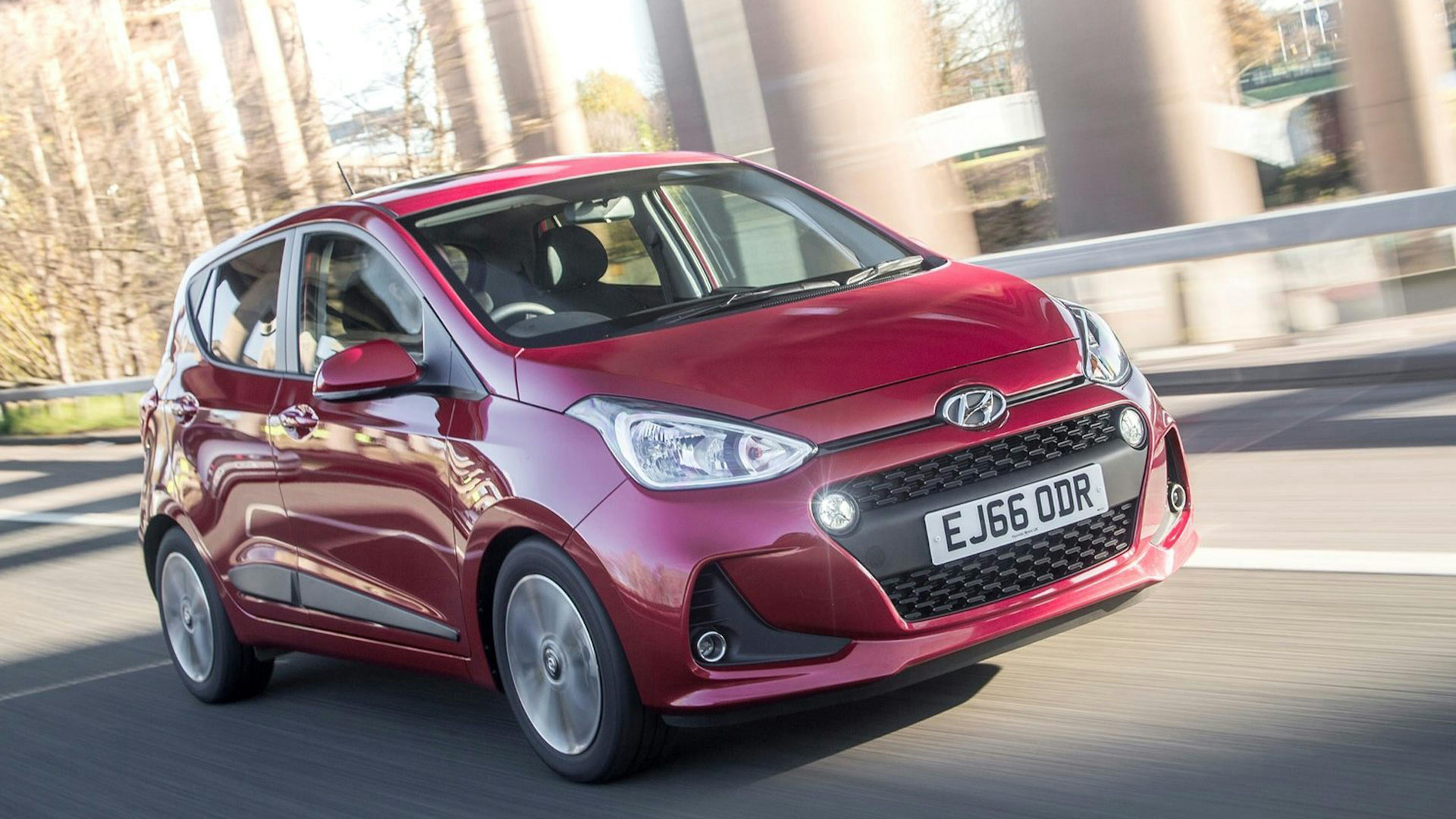 Ein roter Hyundai i10 Kleinstwagen fährt durch eine urbane Kulisse.