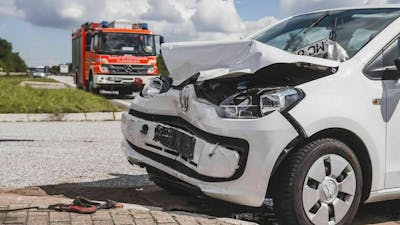 Ein weißer Pkw mit Unfallschaden steht am Straßenrand, im Hintergrund nähert sich ein Feuerwehrauto.