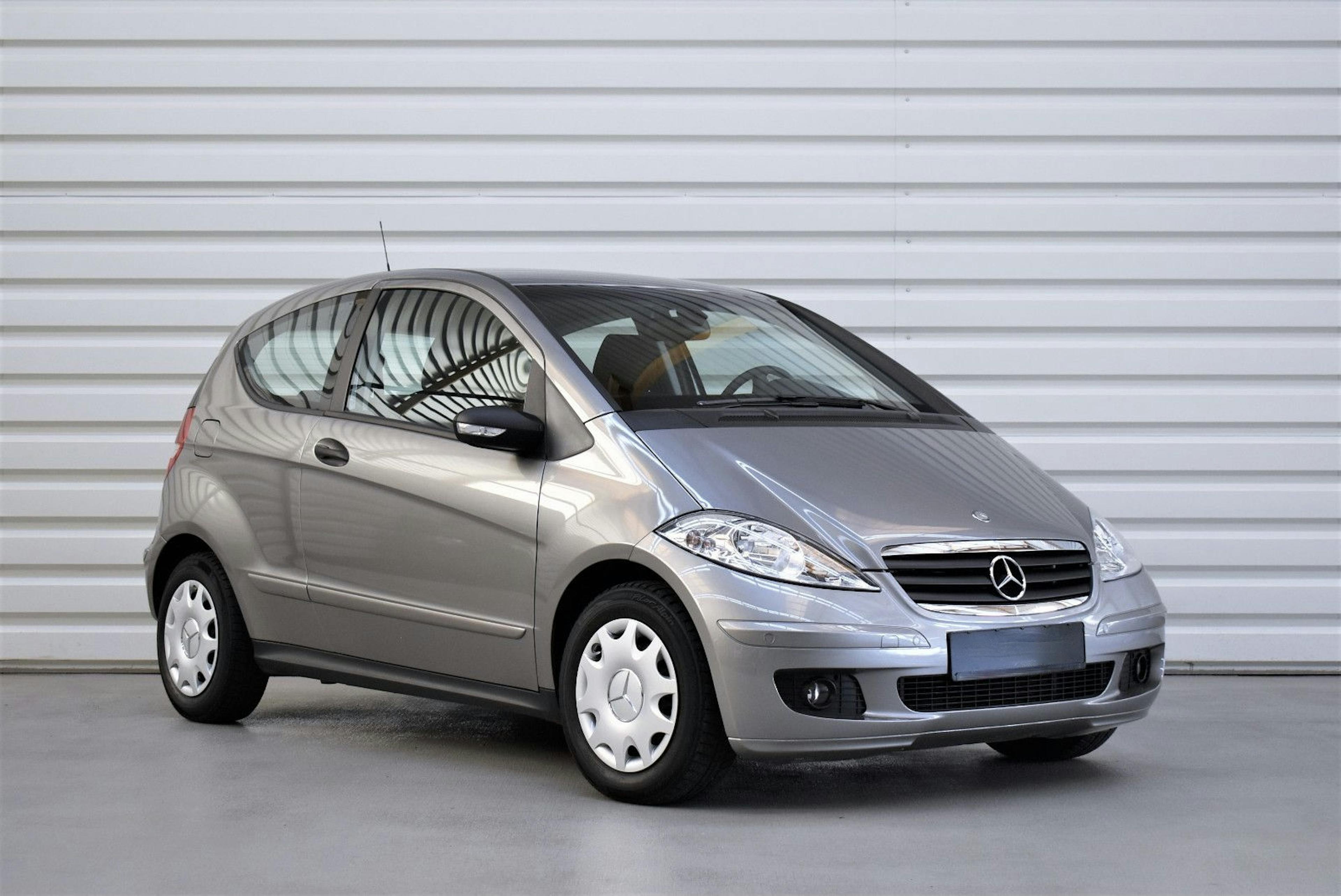 Silberner Mercedes-Benz A-Klasse (W169) steht vor einer weißen Metallwand, leicht seitlich ausgerichtet. Kompakter Stadtwagen mit schlichtem Design.