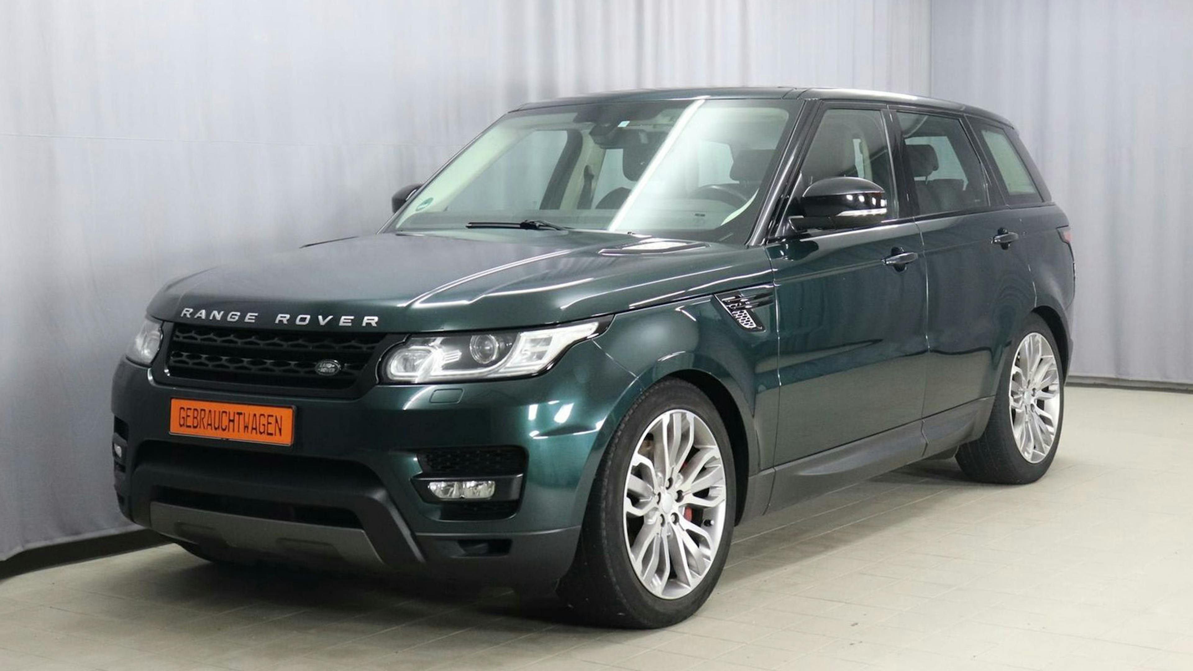 Ein dunkelgrüner Land Rover Range Rover Sport 3.0 225 kW steht vor einem grauen Vorhang.