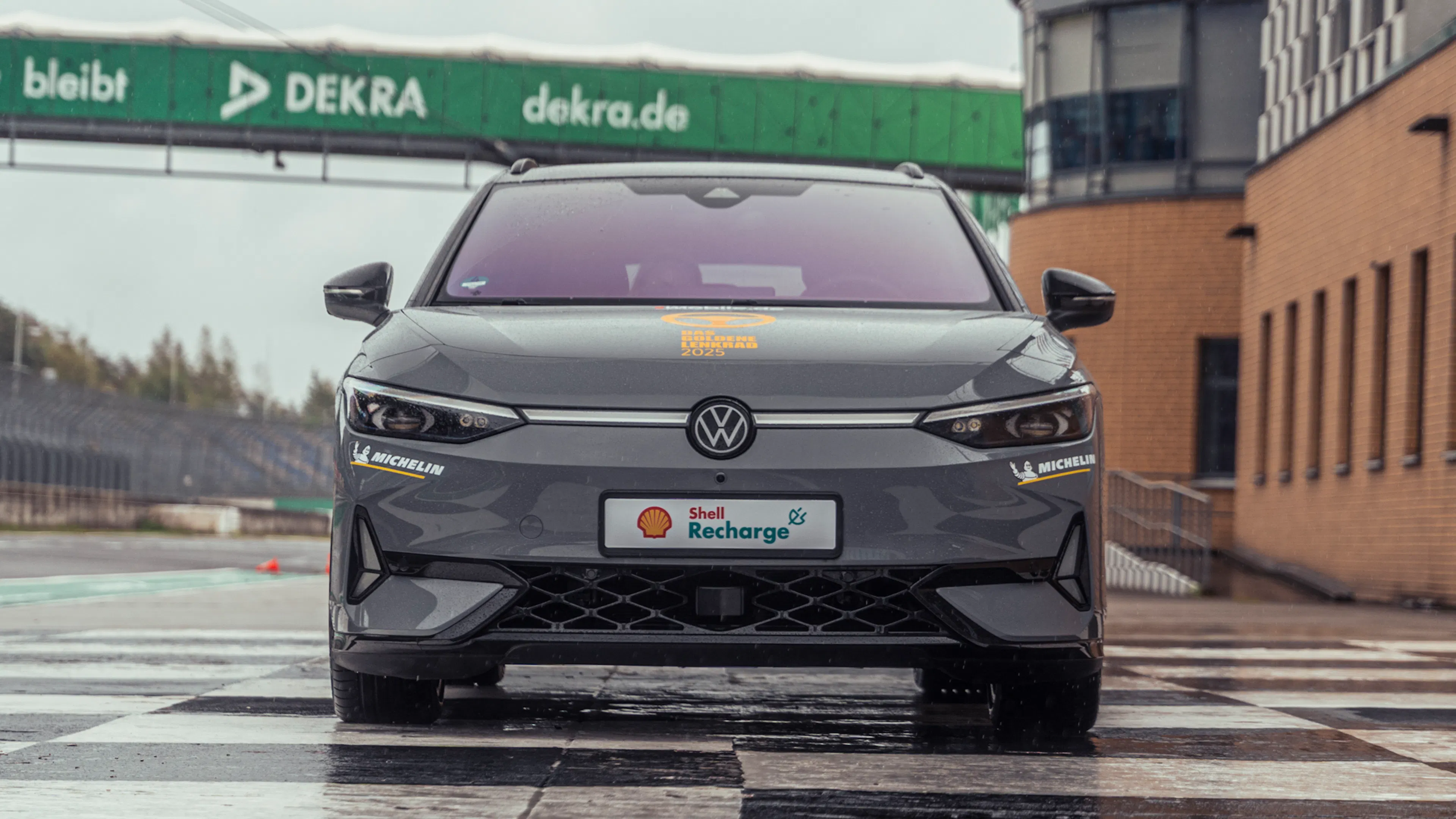 Frontansicht eines grauen VW ID.7 Tourer GTX auf nasser Strecke vor Gebäuden am Lausitzring.