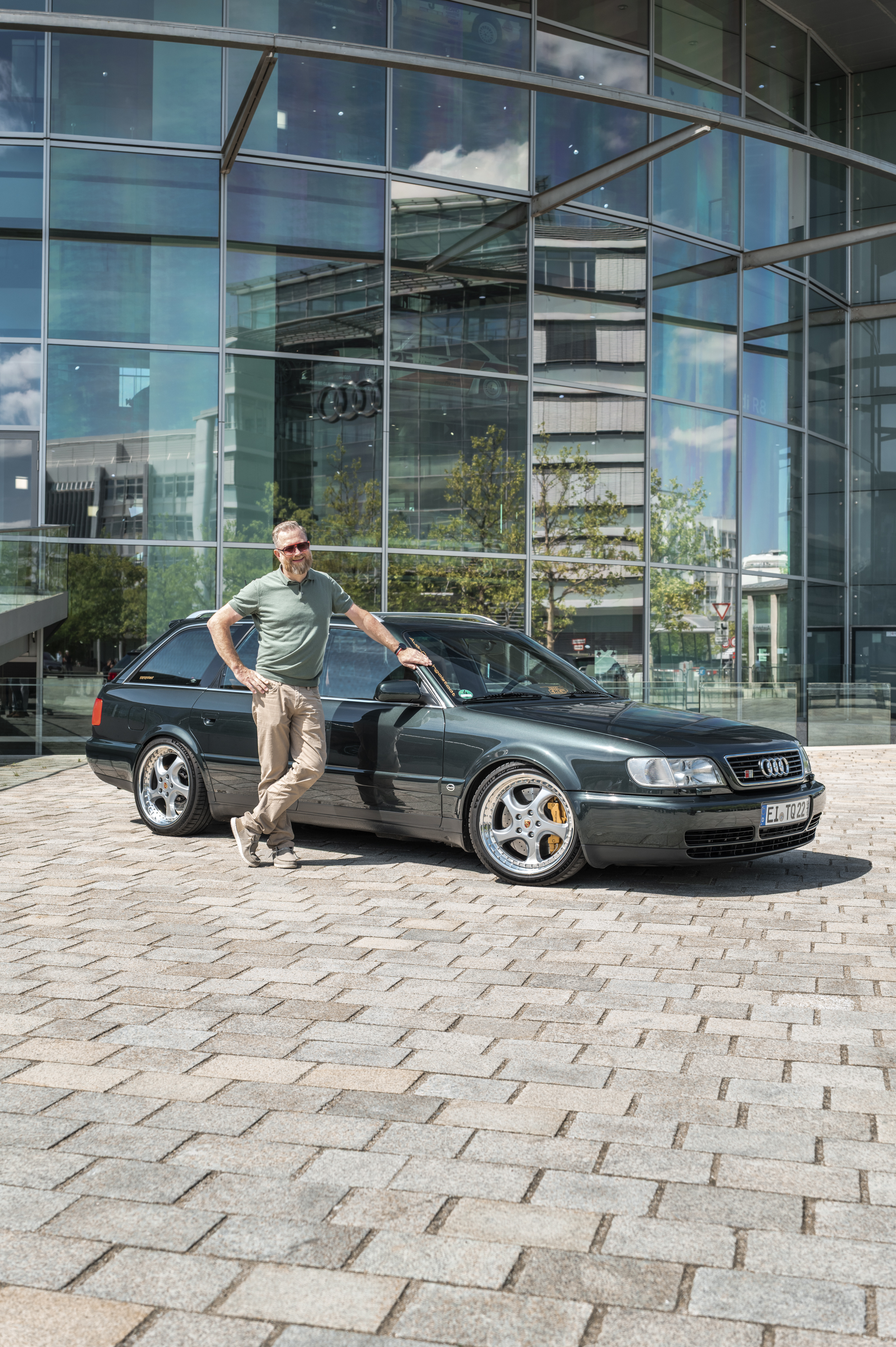 Dunkelgrüner Audi S6 Avant der Generation 100 C4/A6 C4 mit Tieferlegung und Besitzer davor stehend vor einer Glasfassade.