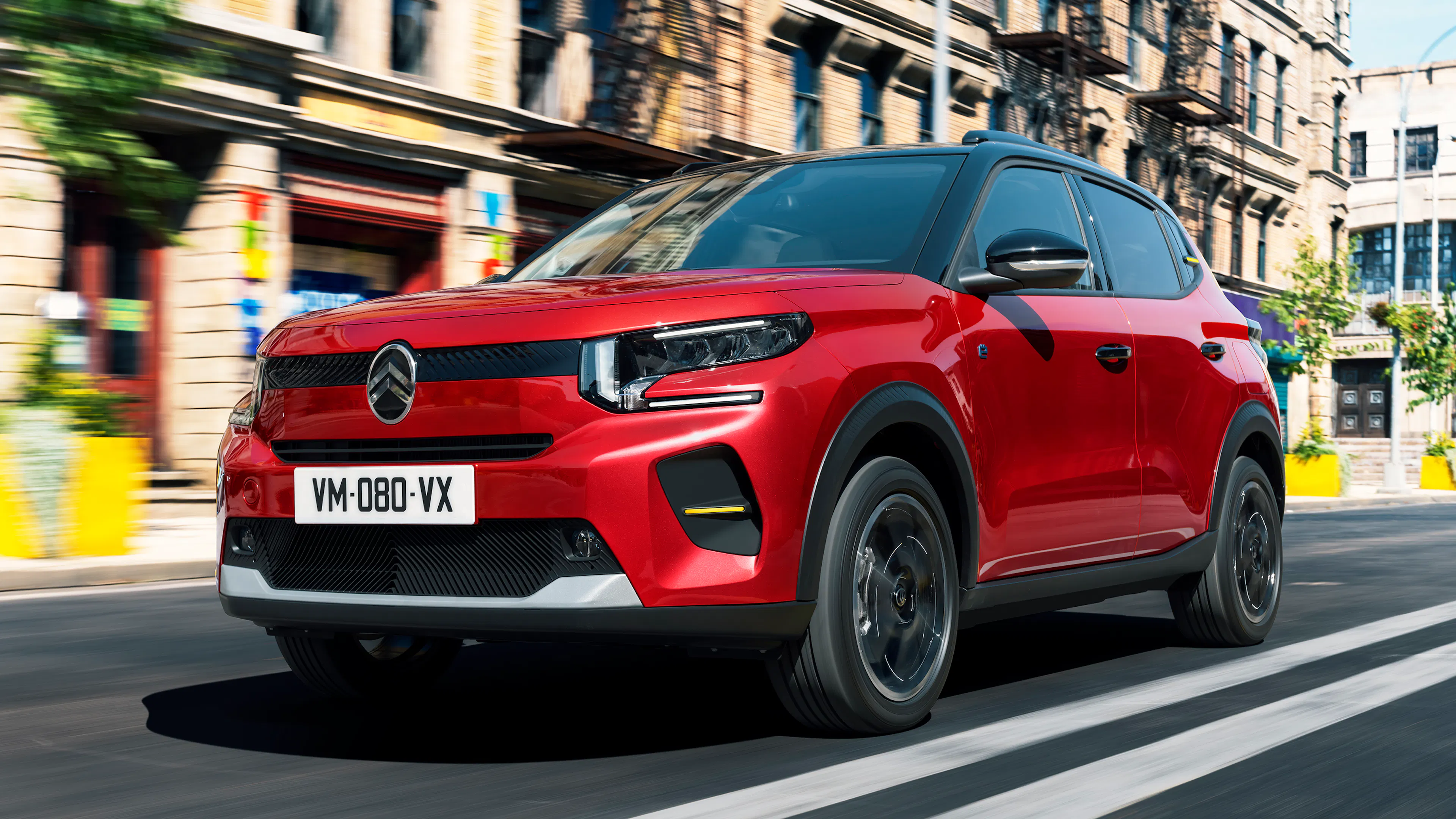 Roter Citroën ë-C3 mit schwarzem Dach und schwarzen Radläufen fährt durch eine Straße mit Altbauten.