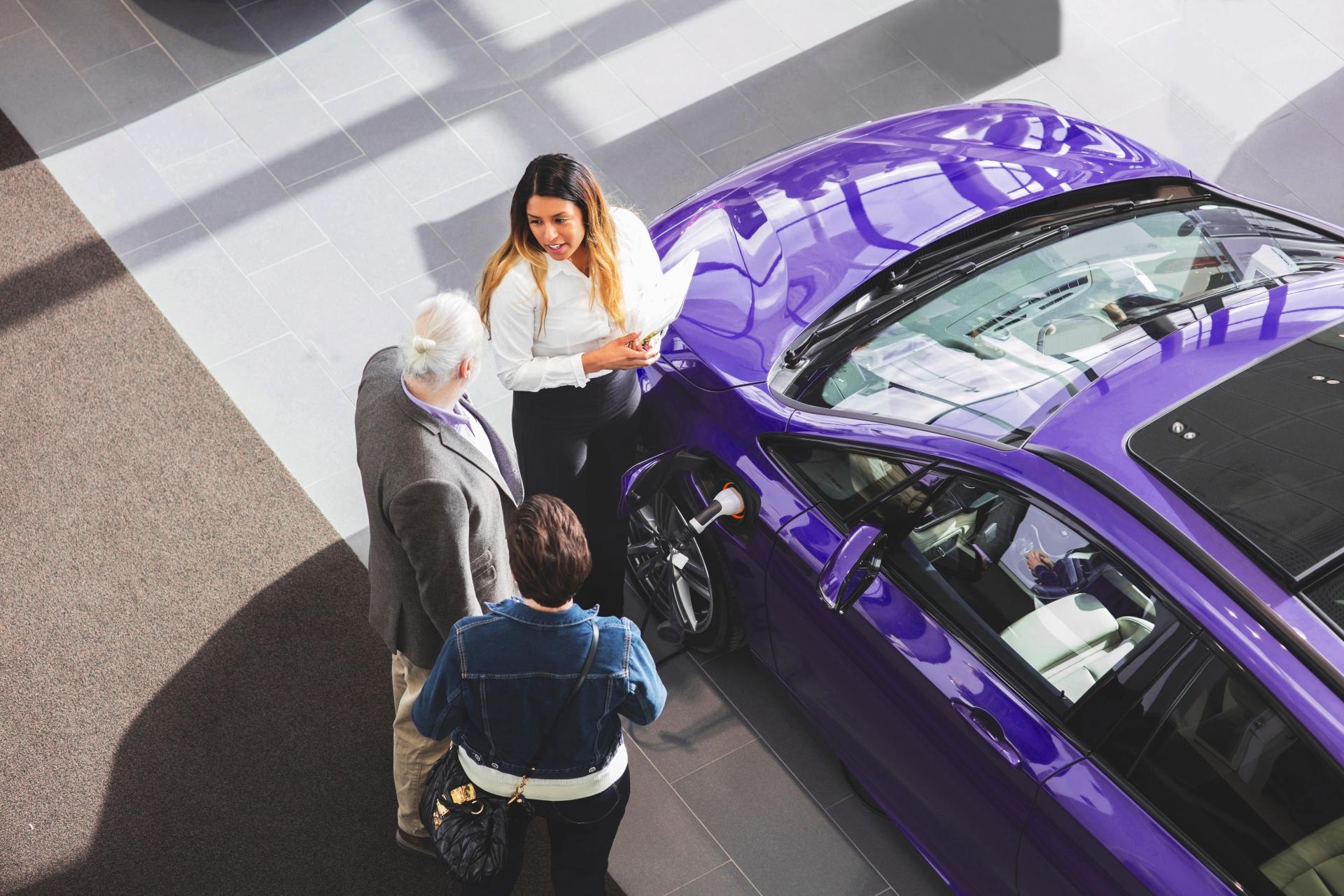 Drei Personen stehen um ein Auto herum. Eine der Frauen möchte das Auto im Kundenauftrag verkaufen.