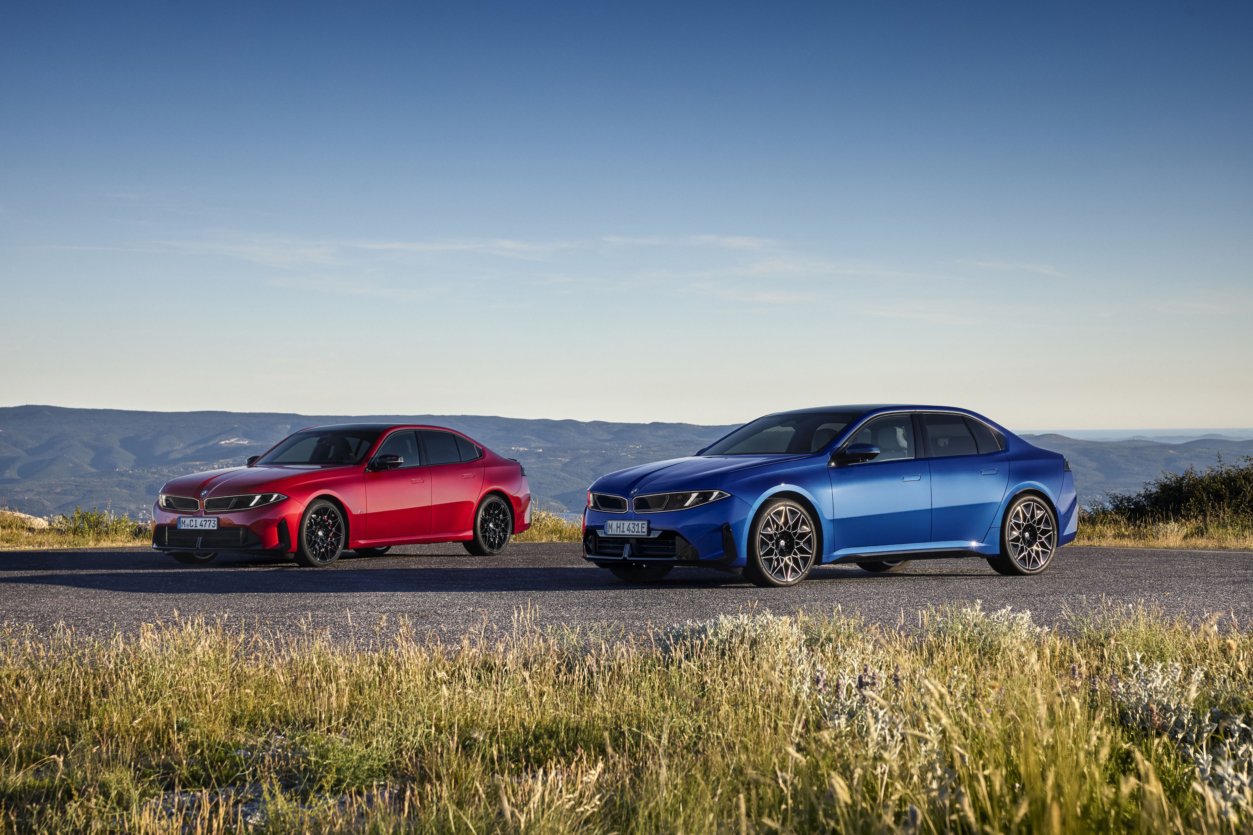 Zwei BMW Limousinen, eine rote und eine blaue, nebeneinander auf einer Bergstraße