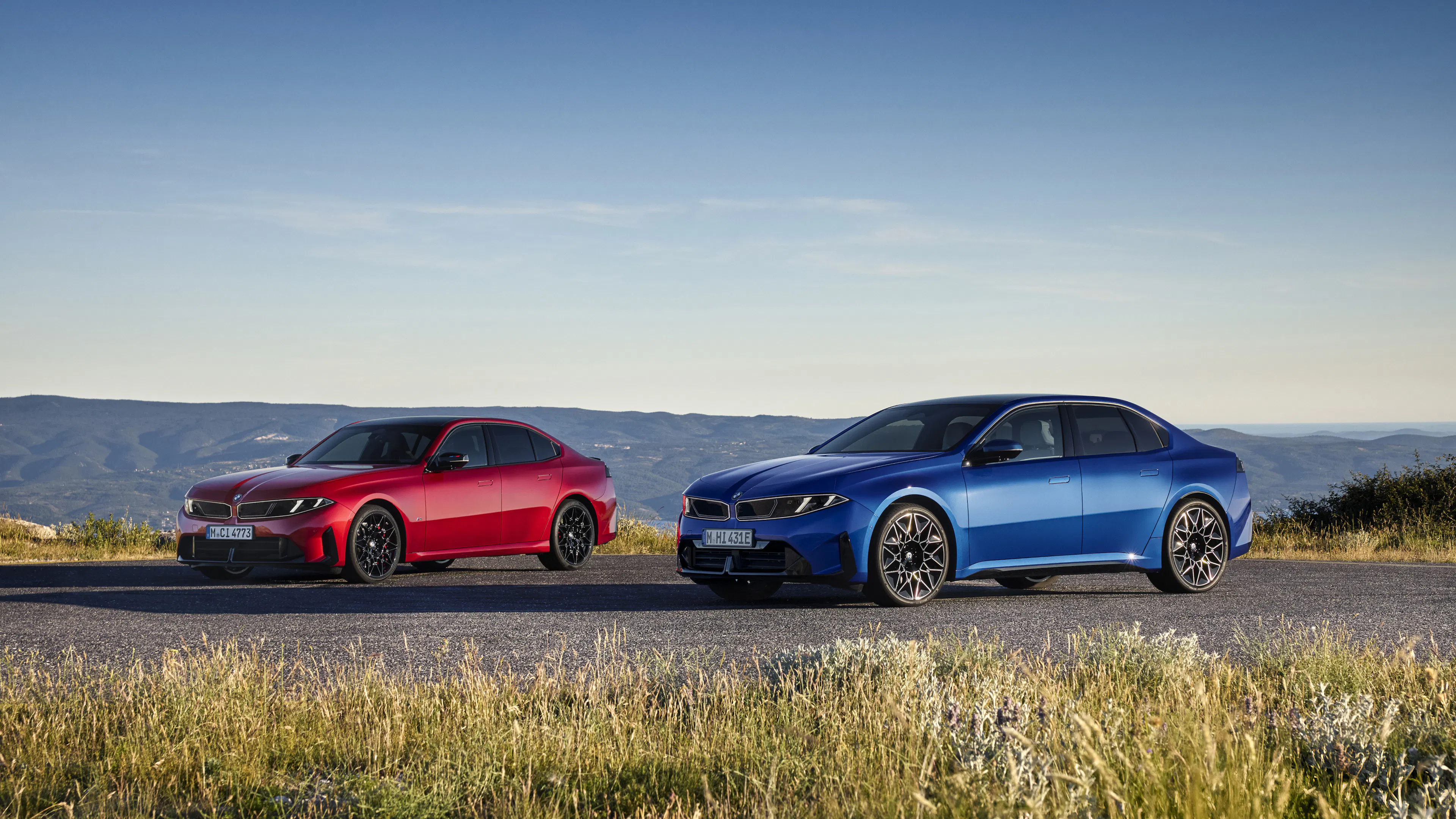 Zwei BMW Limousinen, eine rote und eine blaue, nebeneinander auf einer Bergstraße