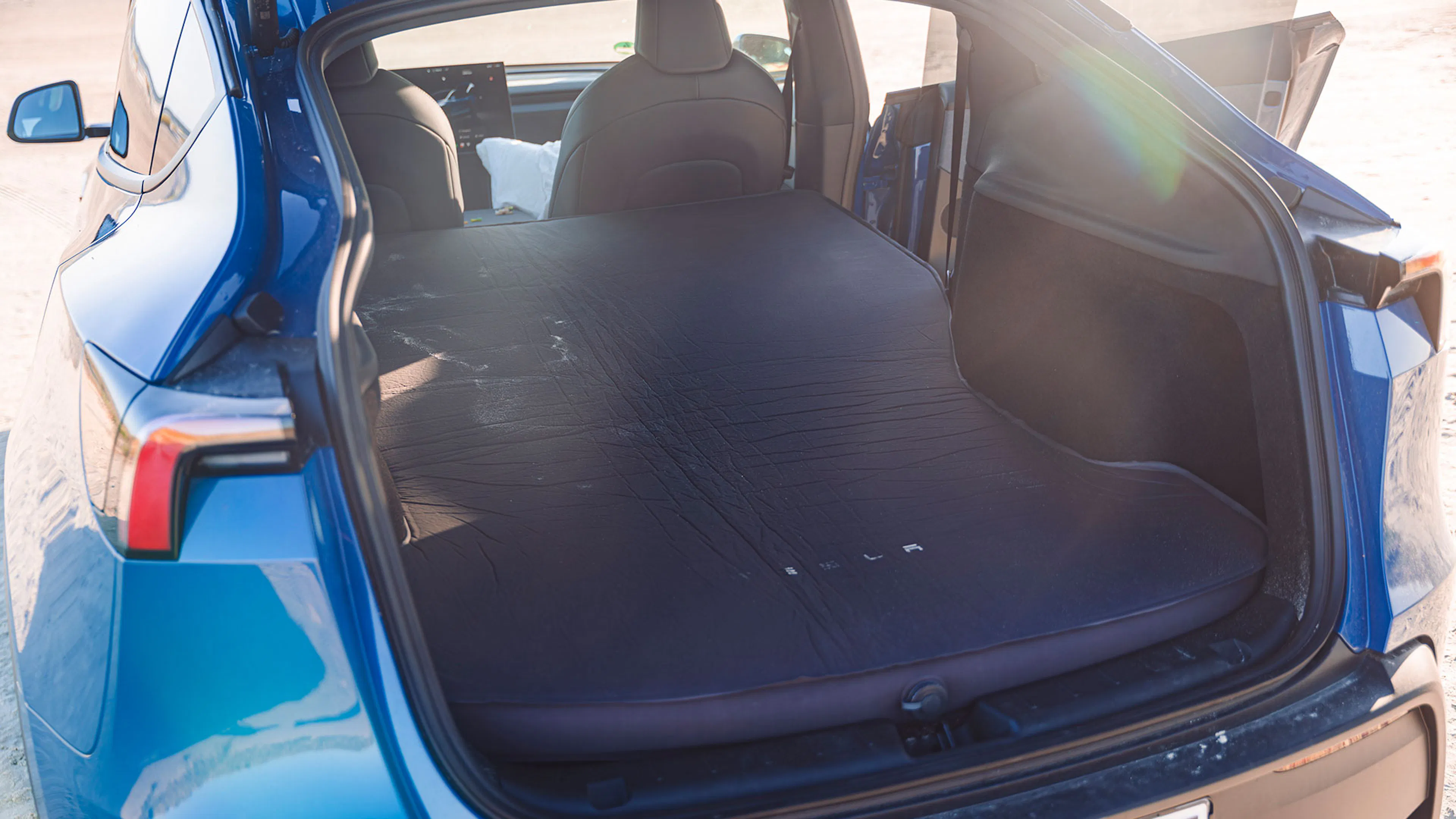 Die Kofferraumklappe eines blauen Tesla Model Y ist geöffnet, die Sitze umgelegt und eine schwarze Luftmatratze aus dem Tesla-Zubehör liegt auf der ebenen Ladefläche.