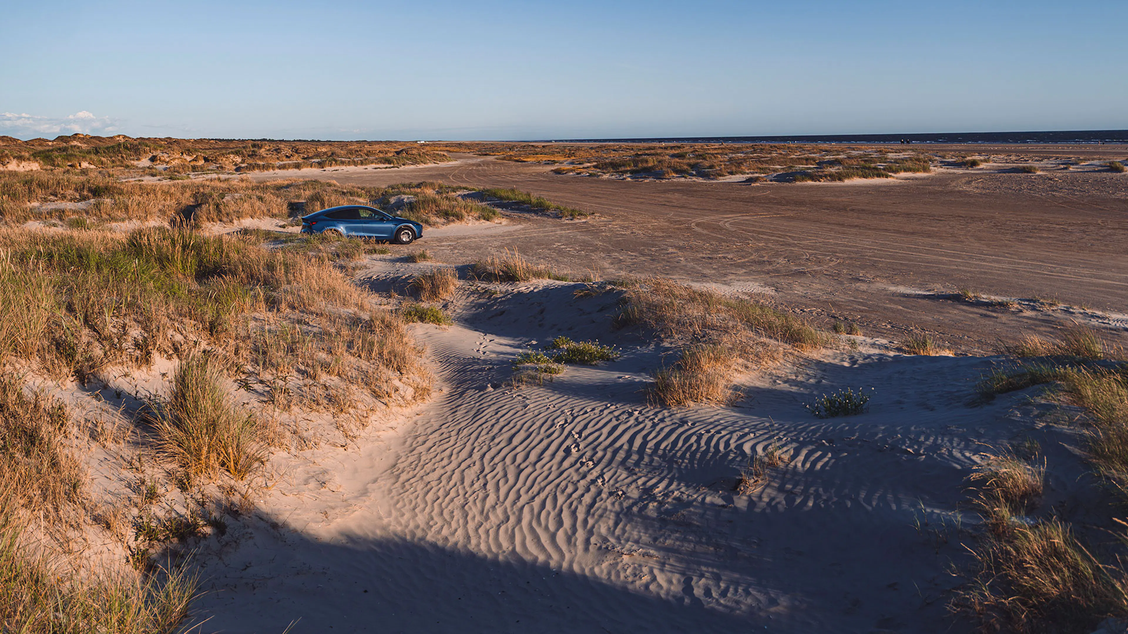 Ein Tesla Model Y steht einsam in einer weitläufigen Dünenlandschaft, im Hintergrund ist das Meer.