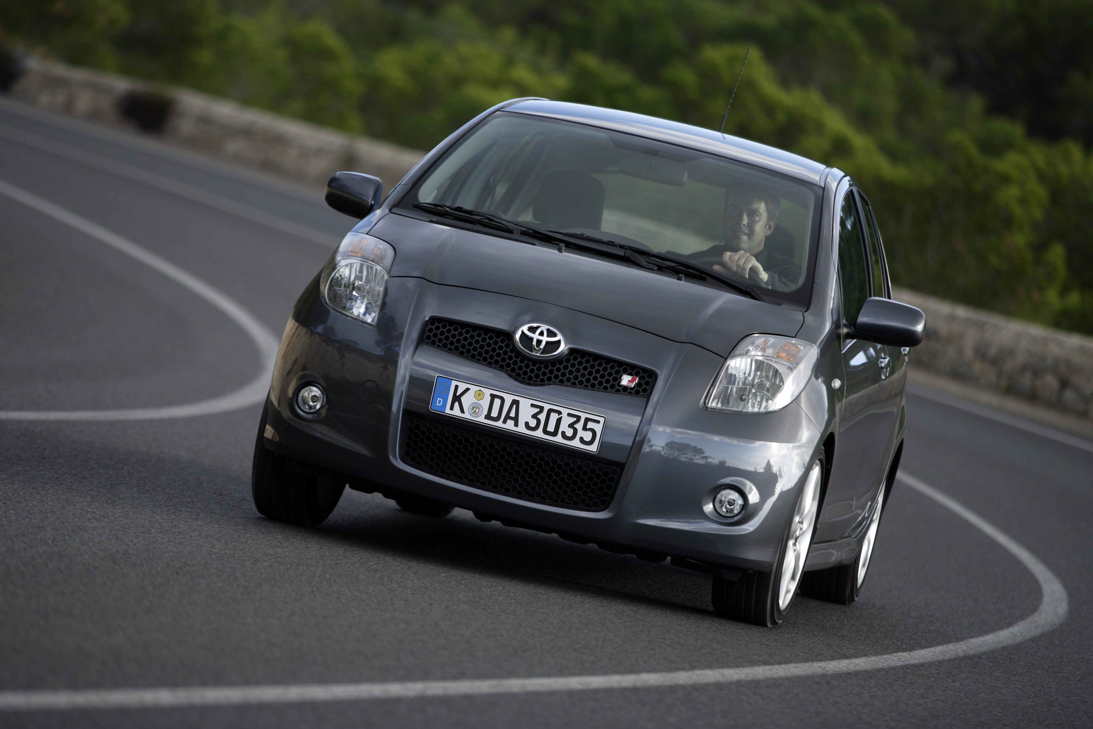 Grauer Toyota Yaris TS fährt frontal auf einer kurvigen Landstraße, am Steuer sitzt ein lächelnder Fahrer.