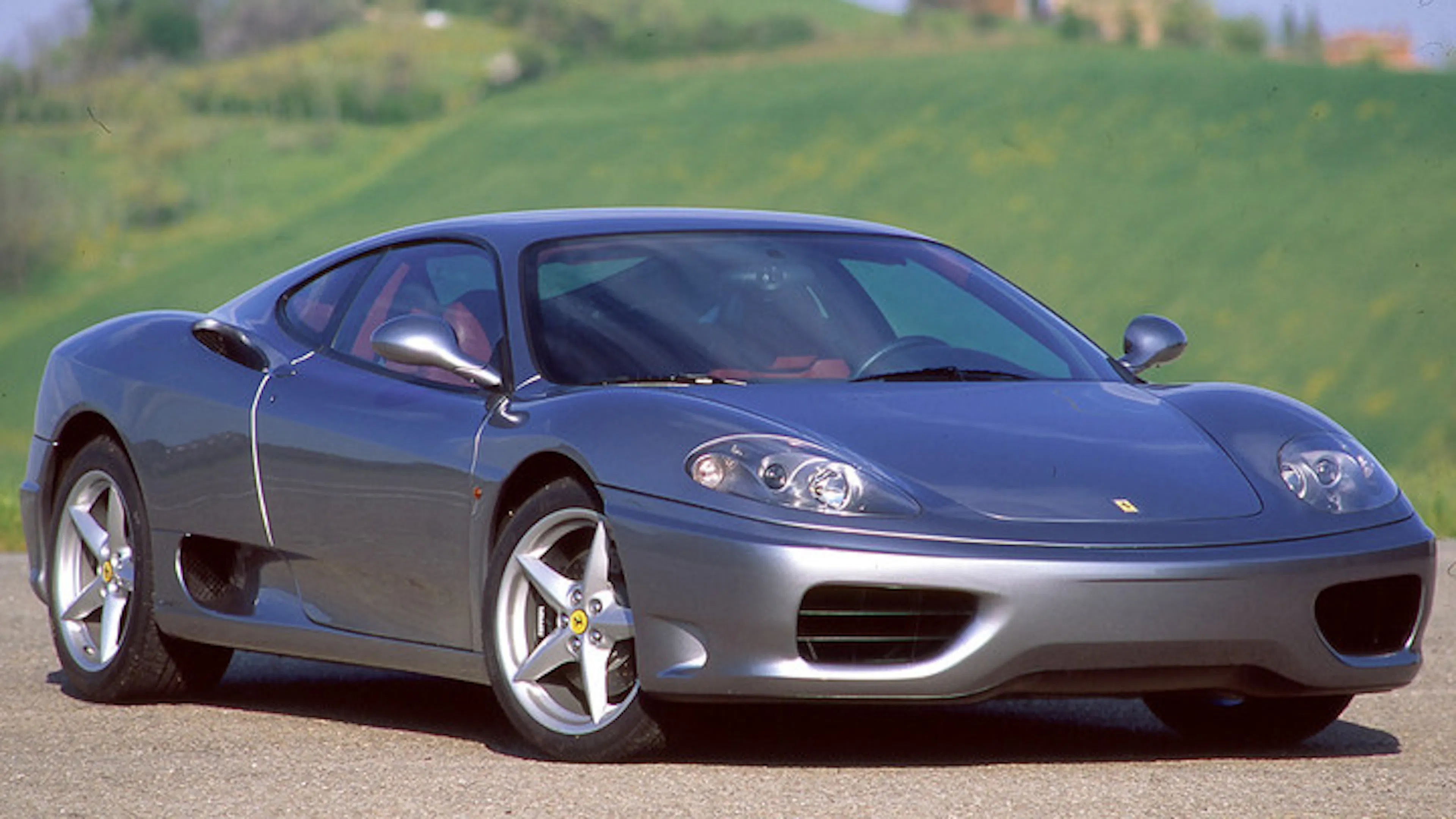 Ein silberner Ferrari 360 Modena steht vor einem grünen Hügel.