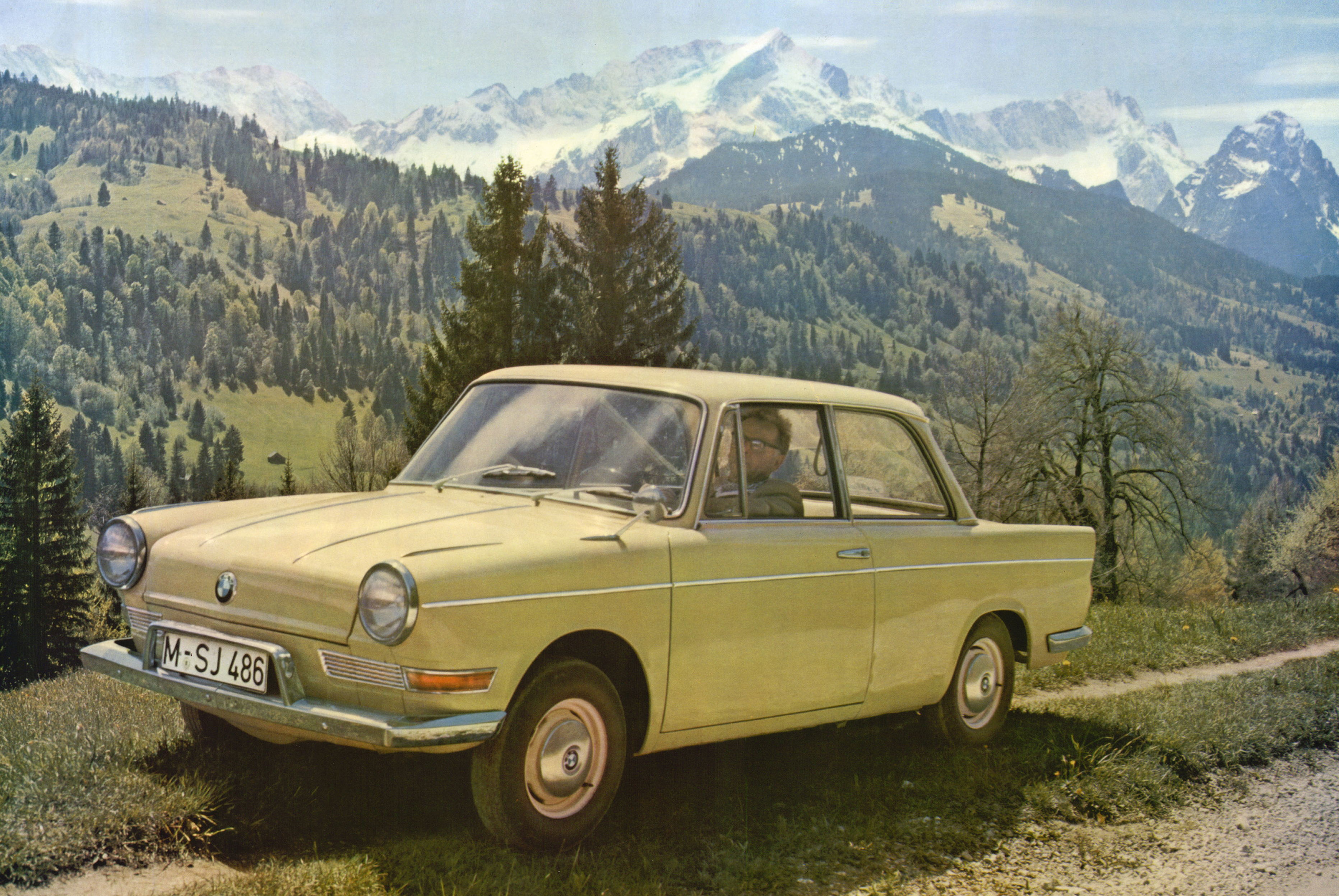 Beigefarbener BMW 700 in der Seitenansicht auf einer Bergstraße vor verschneiten Alpengipfeln.