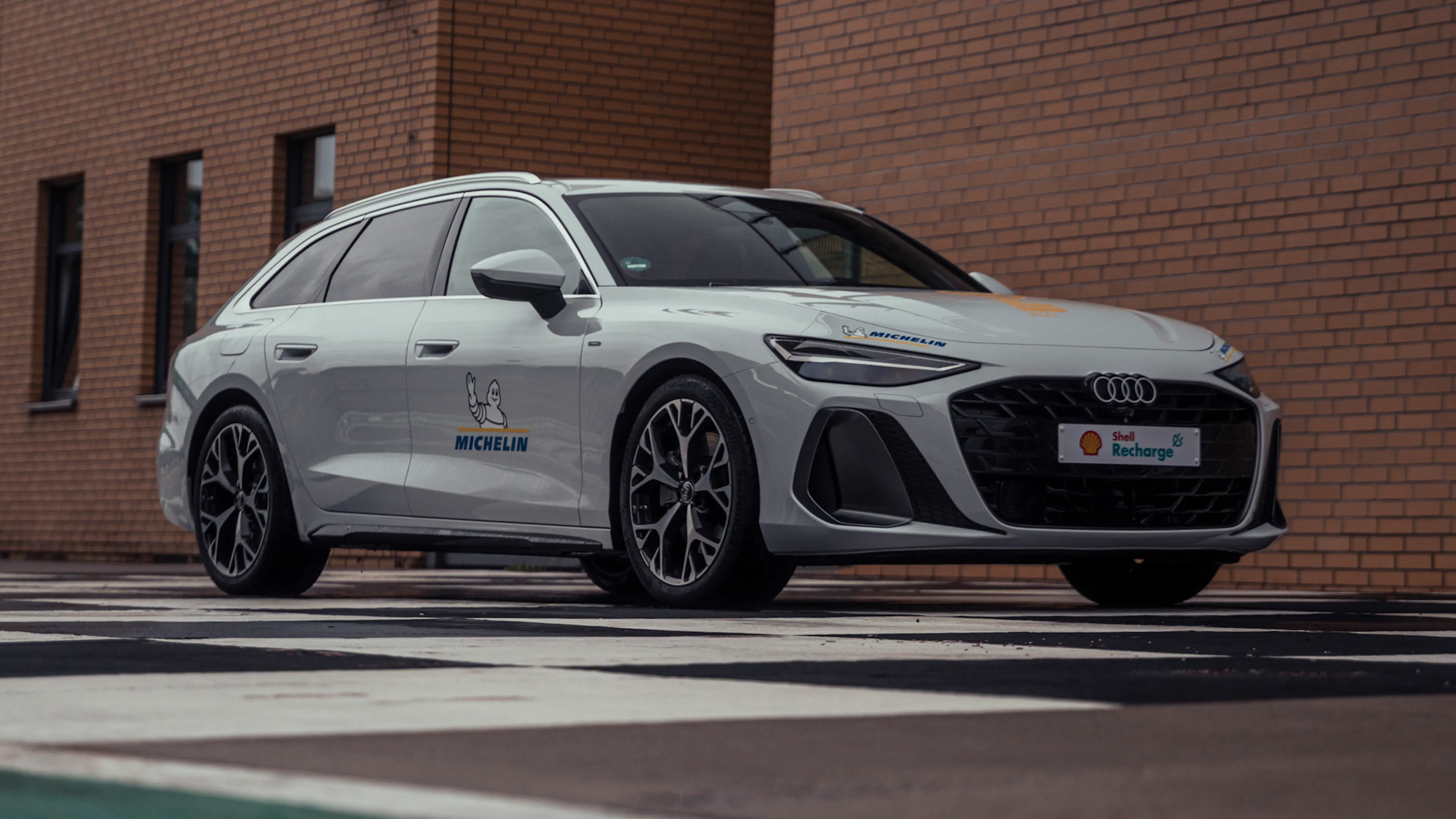 Weißer Audi A6 Avant Plug-in-Hybrid in Schrägansicht von vorn vor einer Backsteinfassade.