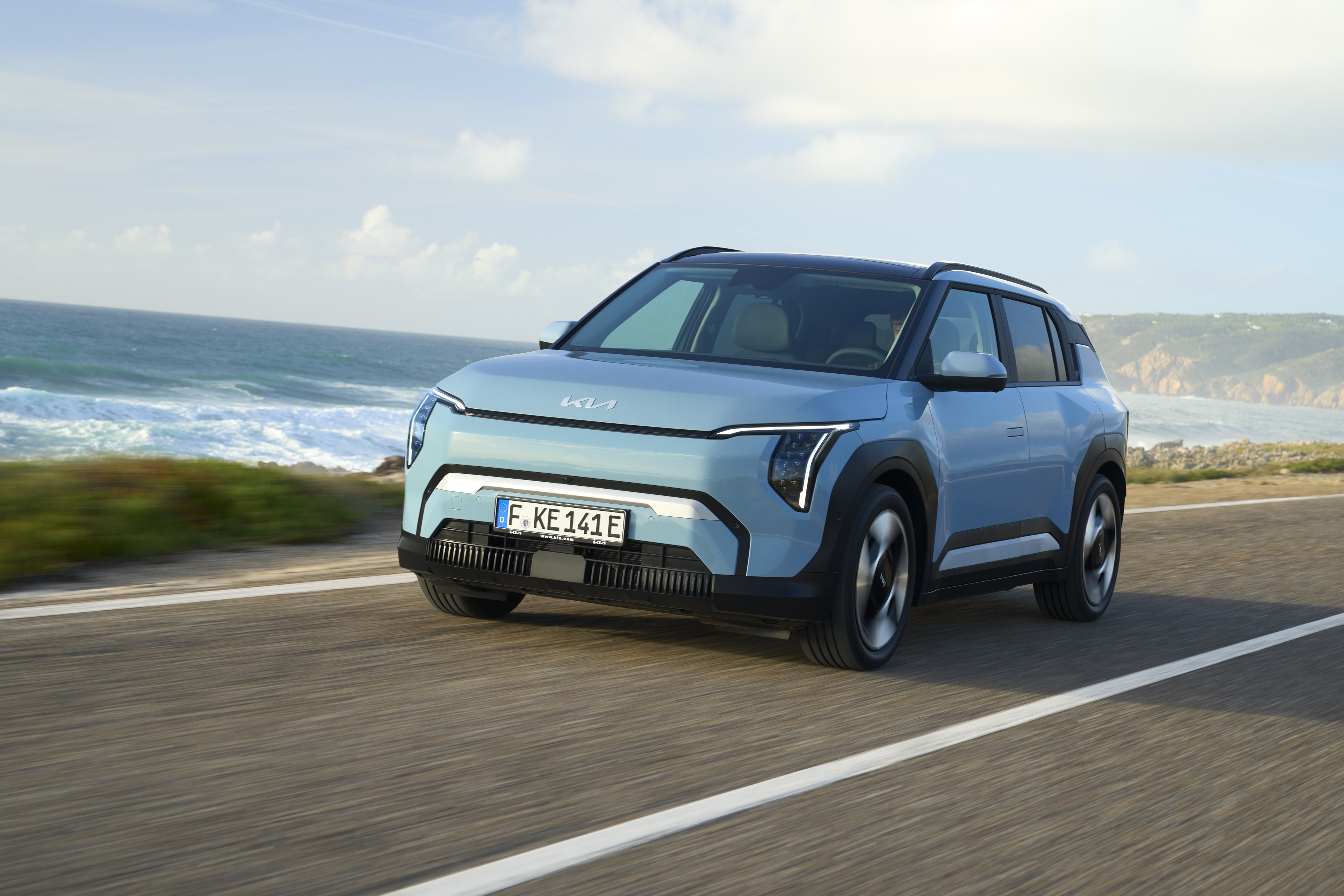 Hellblauer Kia EV3 fährt schräg von vorn gesehen auf einer Küstenstraße, dahinter das Meer.