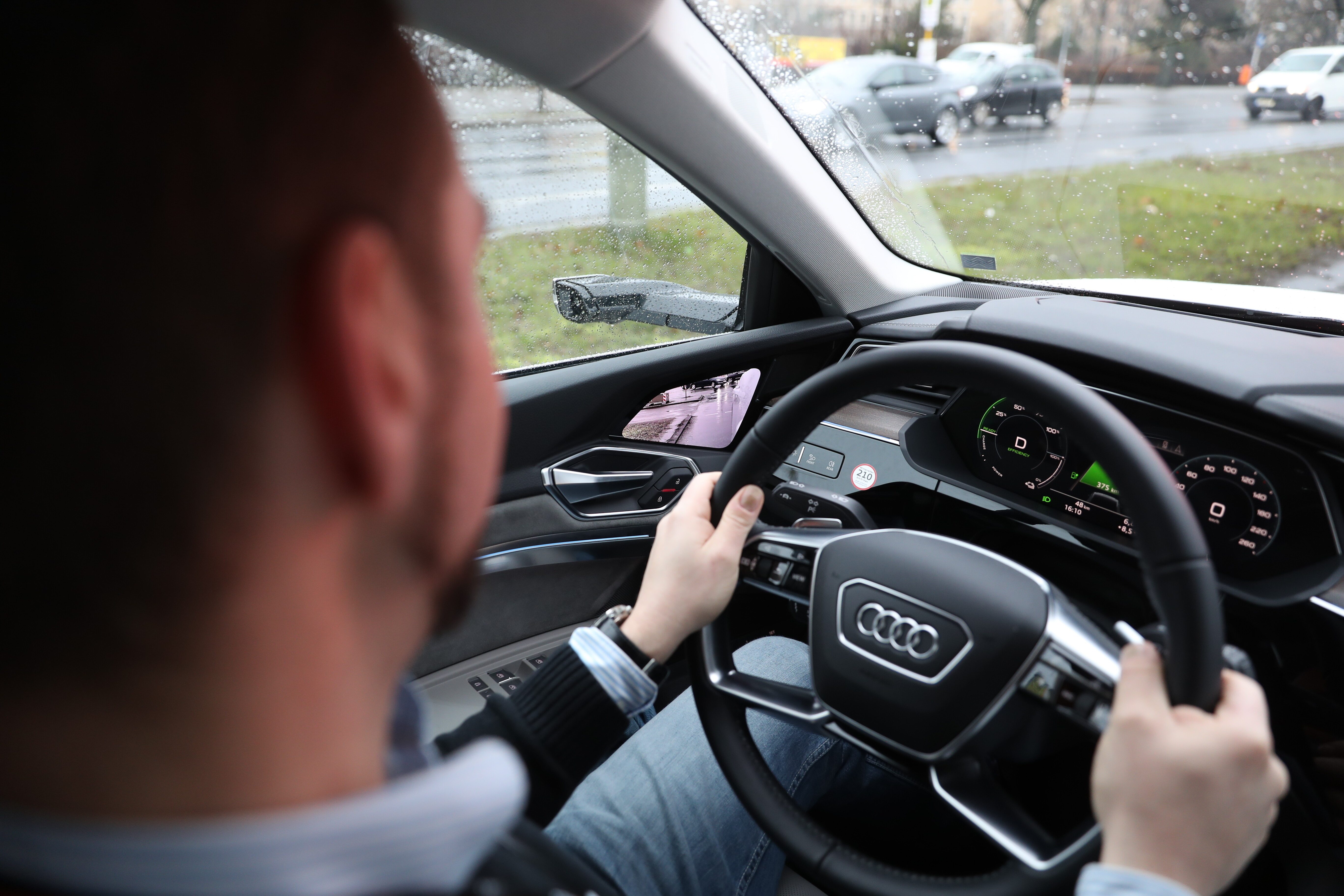 Fahrer hält beim Probefahren mit beiden Händen das Lenkrad eines Audi bei Regen, im Display sind das digitale Cockpit und das Kamerabild des Außenspiegels zu sehen.
