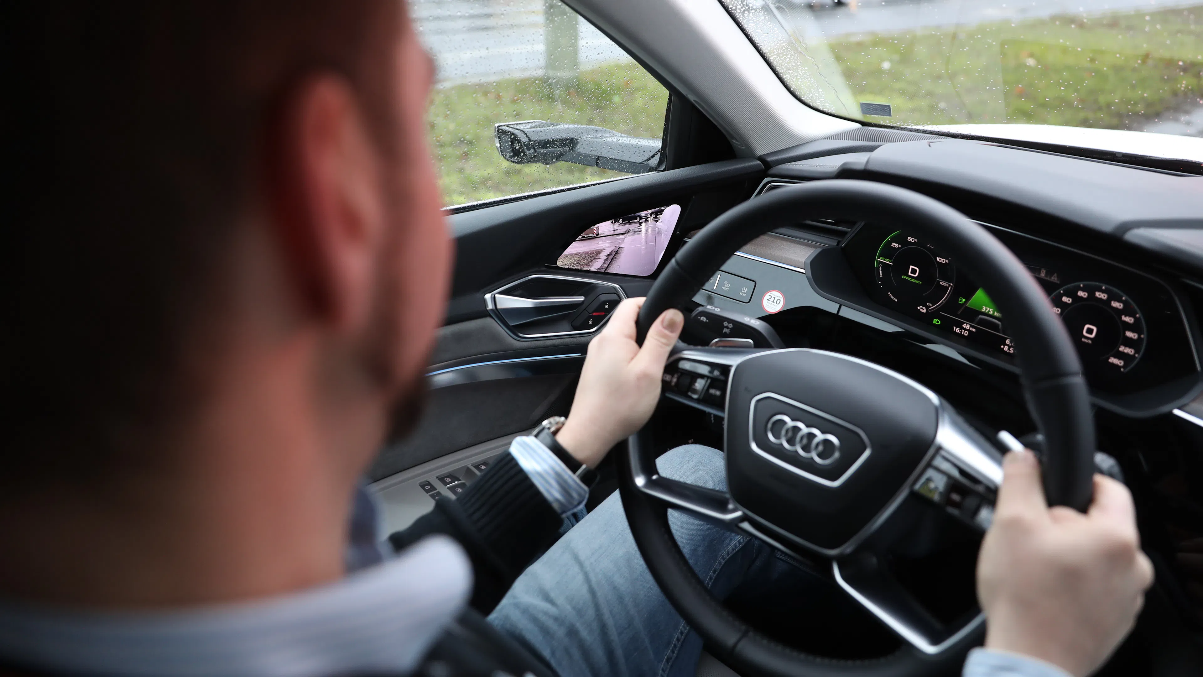 Fahrer hält beim Probefahren mit beiden Händen das Lenkrad eines Audi bei Regen, im Display sind das digitale Cockpit und das Kamerabild des Außenspiegels zu sehen.