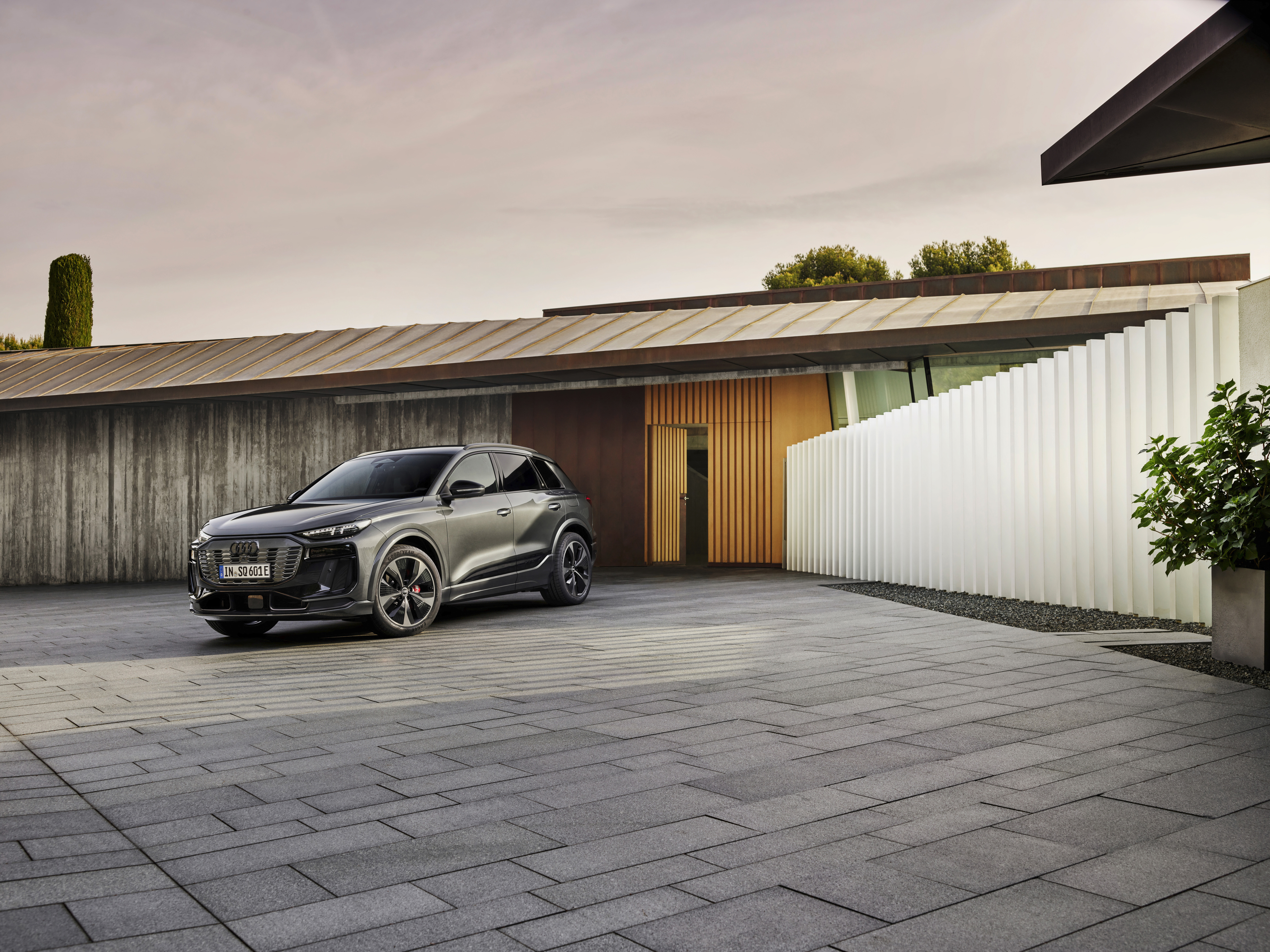 Grauer Audi SQ6 e-tron parkt auf einem gepflasterten Hof vor einem flachen Haus mit Holz- und Betonwänden.