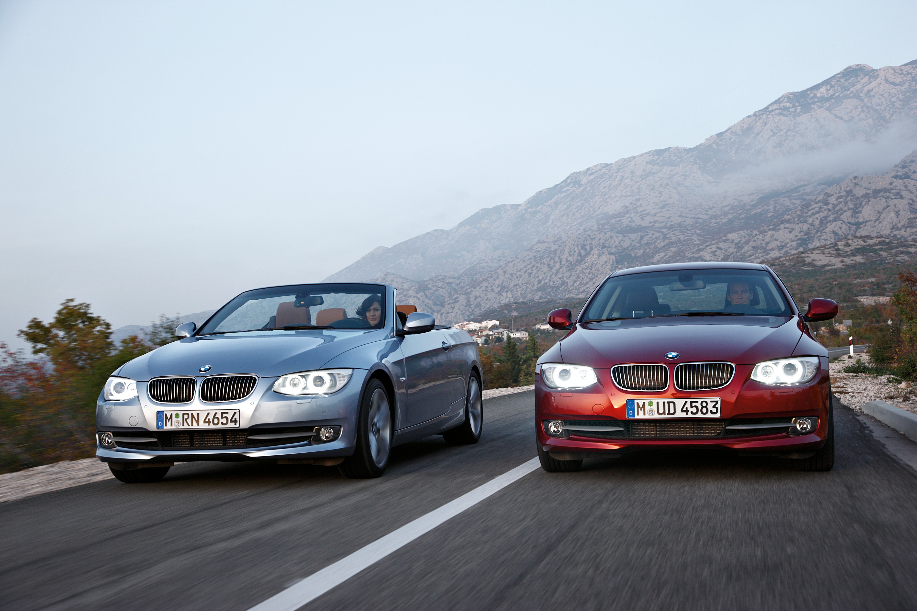 BMW 3er E92 Coupé in Silber und BMW 3er E93 Cabriolet in Dunkelrot fahren nebeneinander auf einer Bergstraße.