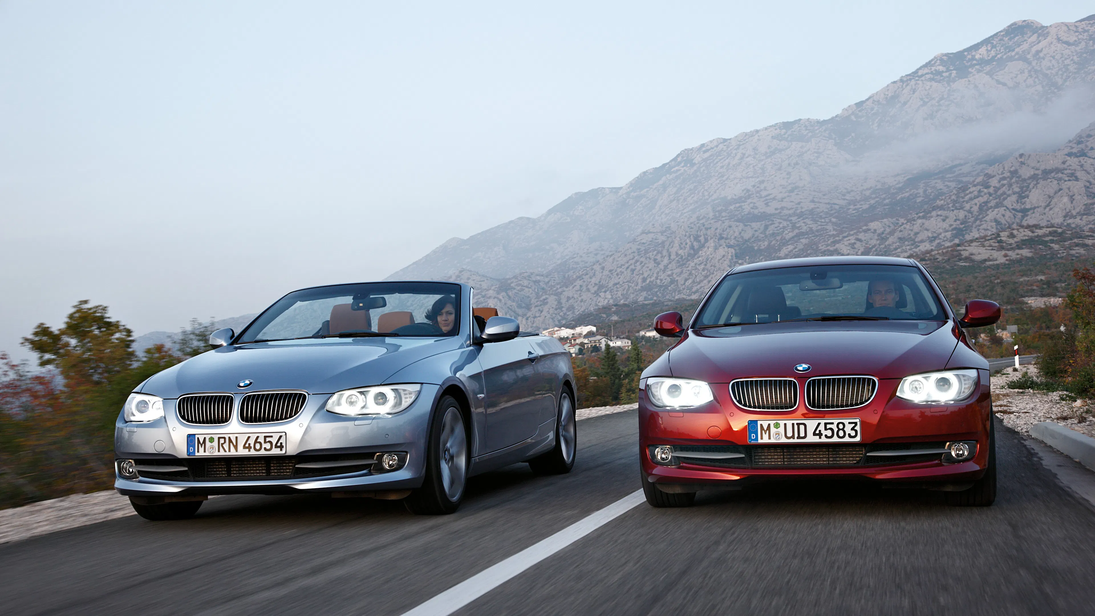 BMW 3er E92 Coupé in Silber und BMW 3er E93 Cabriolet in Dunkelrot fahren nebeneinander auf einer Bergstraße.