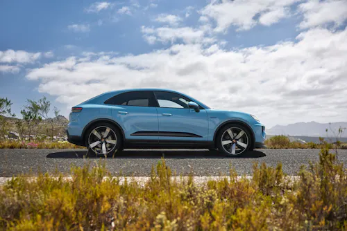 Hellblauer Porsche Macan Elektro in der Seitenansicht, parkend auf einer Straße vor einer Berglandschaft mit blauem Himmel und Pflanzen im Vordergrund.