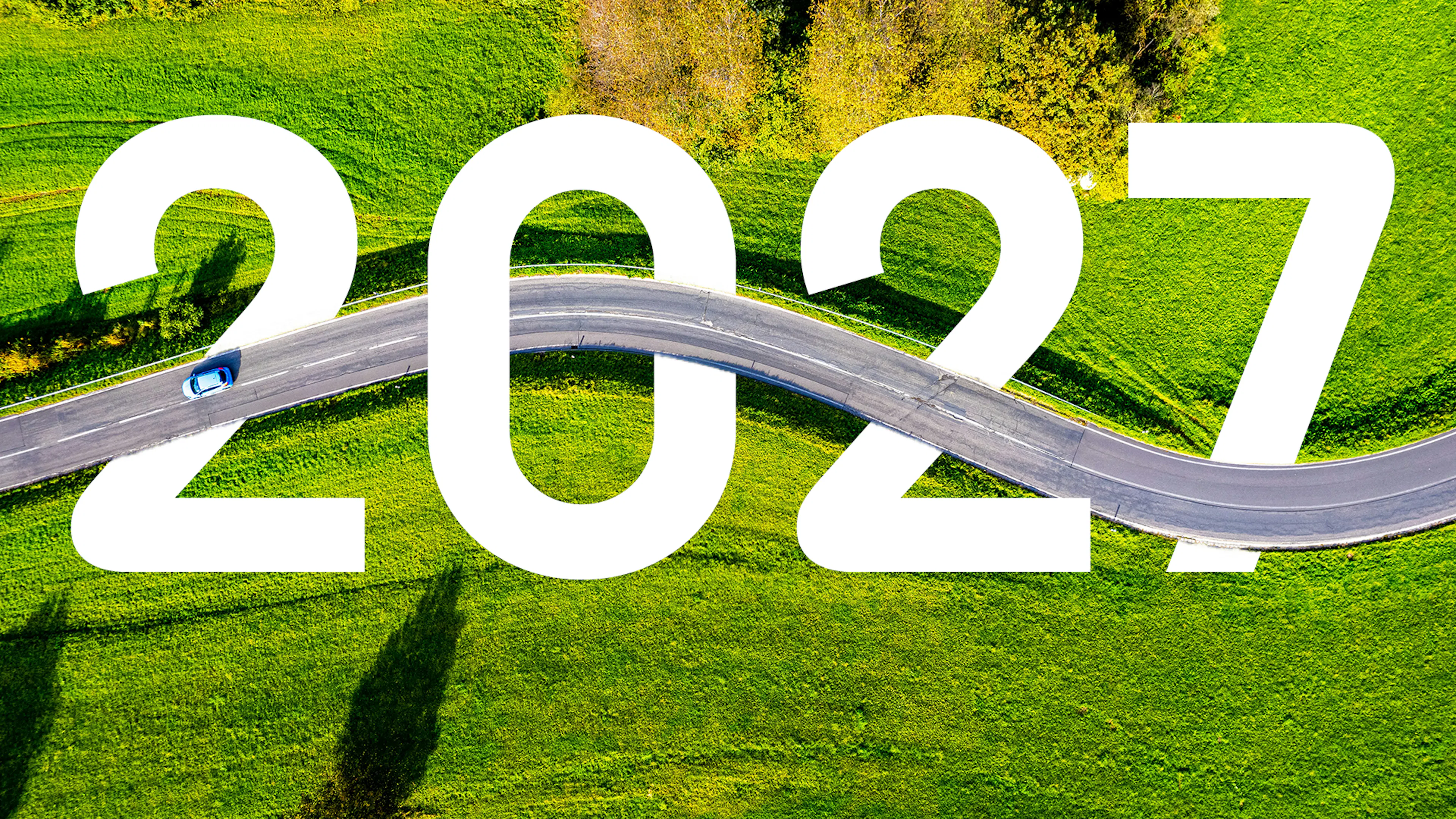 Luftaufnahme einer Landstraße, die sich durch grüne Wiesen schlängelt, mit einem kleinen blauen Auto und der großen weißen Zahl 2027 im Vordergrund