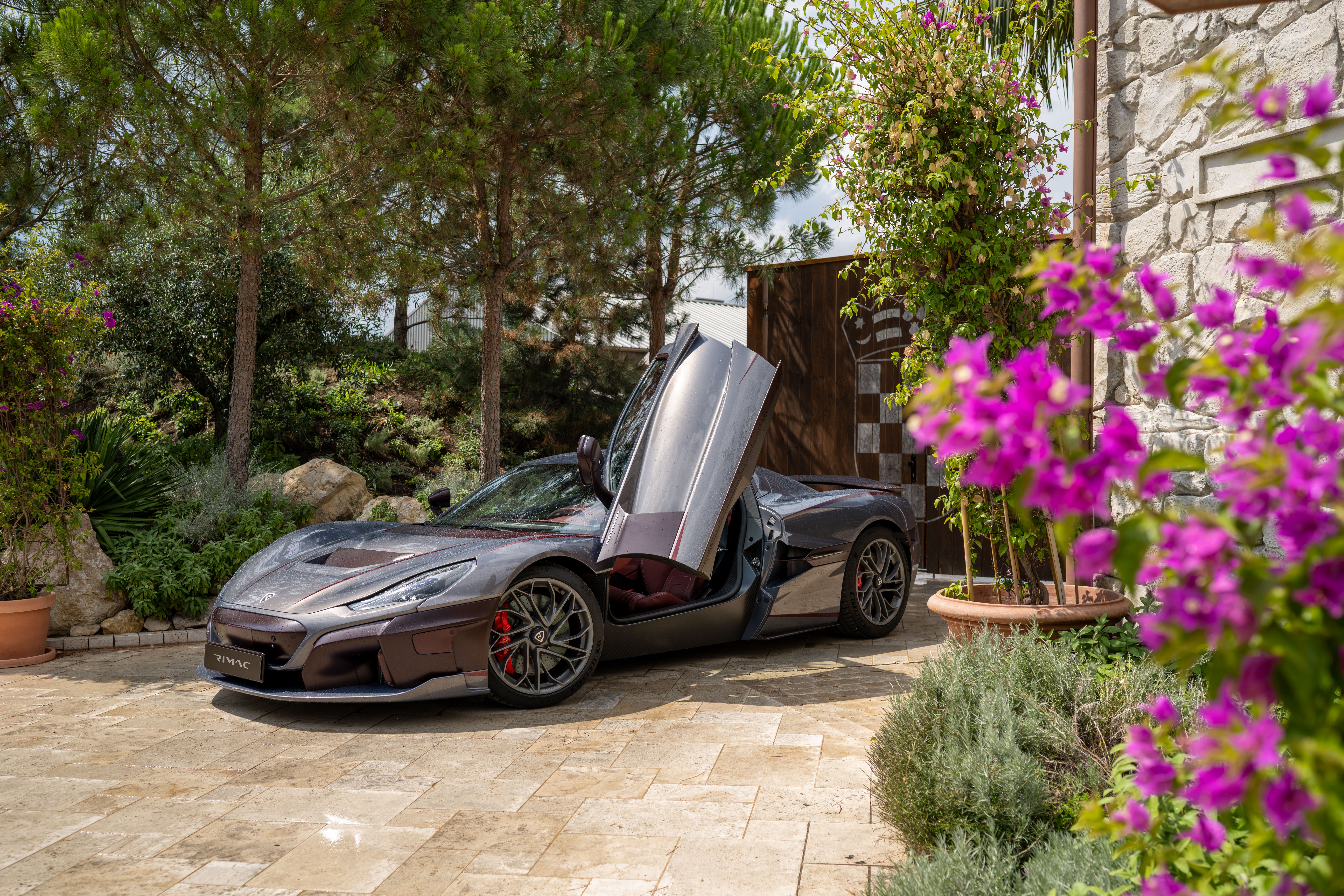 Rimac Nevera in Dunkelbraun mit geöffneten Scherentüren, parkend vor mediterraner Kulisse mit Bougainvillea.