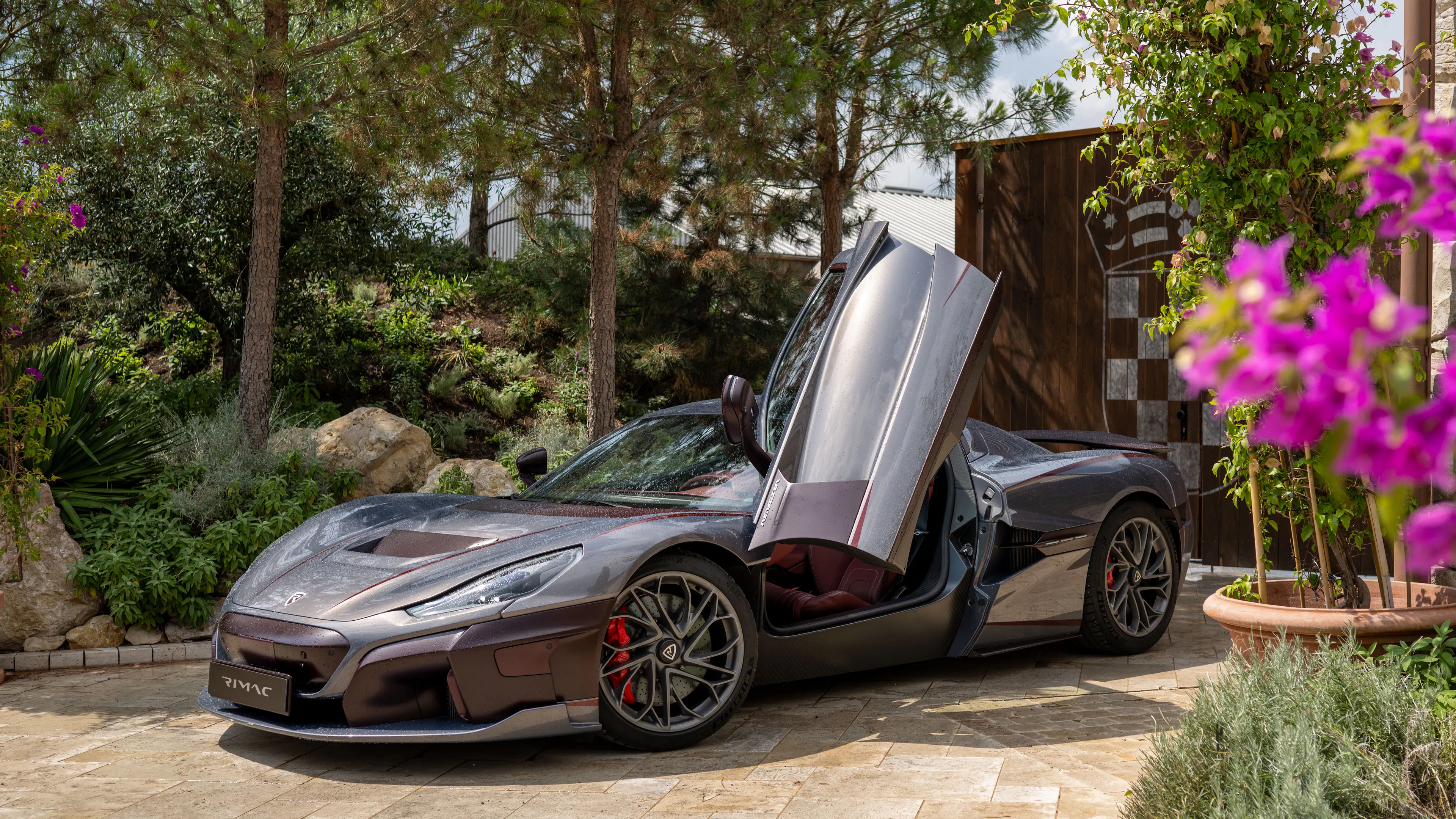 Rimac Nevera in Dunkelbraun mit geöffneten Scherentüren, parkend vor mediterraner Kulisse mit Bougainvillea.