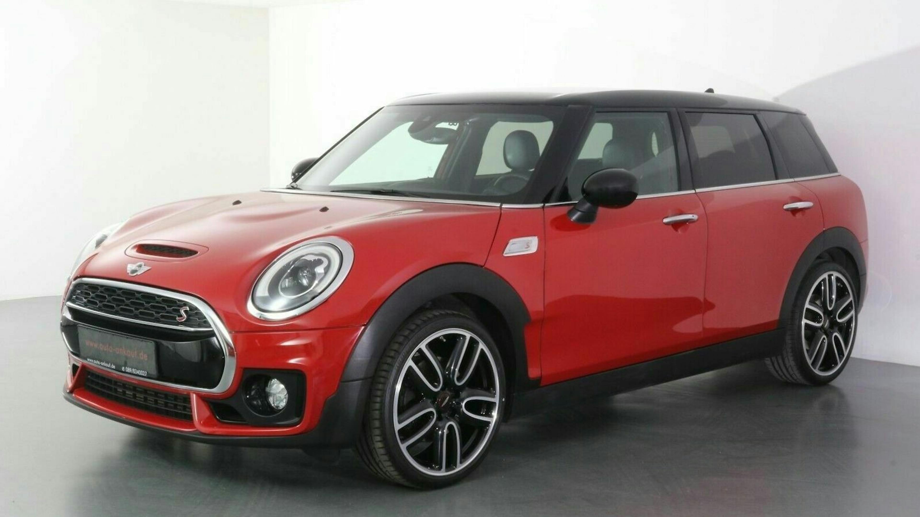 Ein roter Mini Cooper Clubman mit schwarzem Dach steht in einem weißen Studio, mit modernen Felgen und markanten Leuchten.