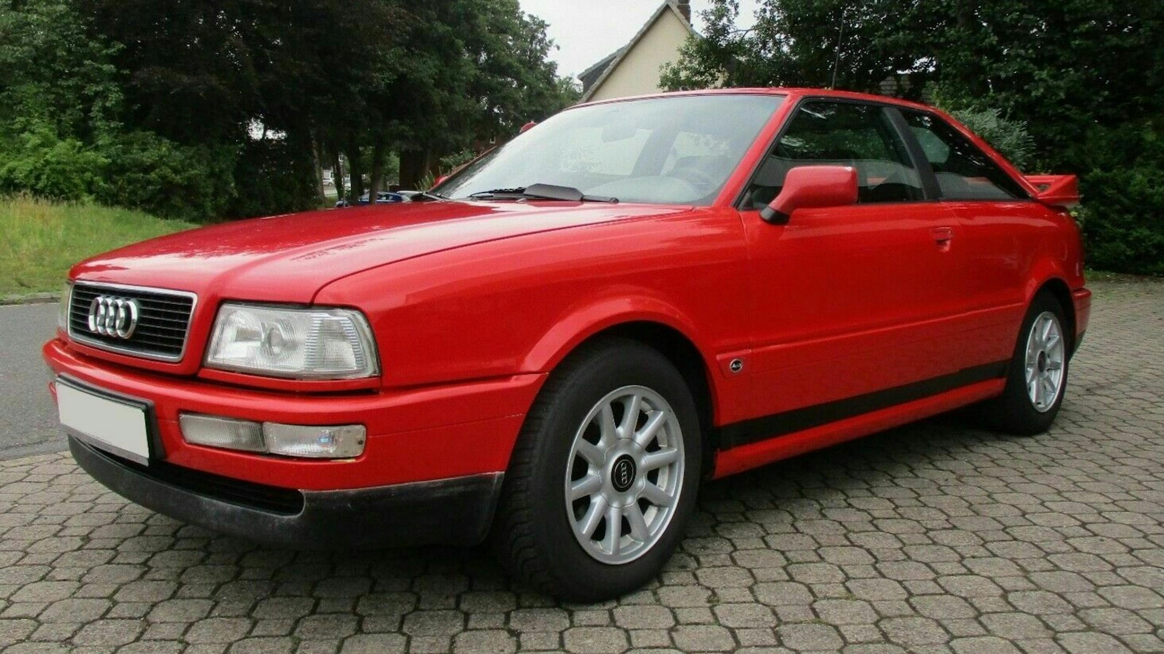 Rotes Audi-Coupé auf gepflasterter Straße, umgeben von Bäumen und einem Haus im Hintergrund.