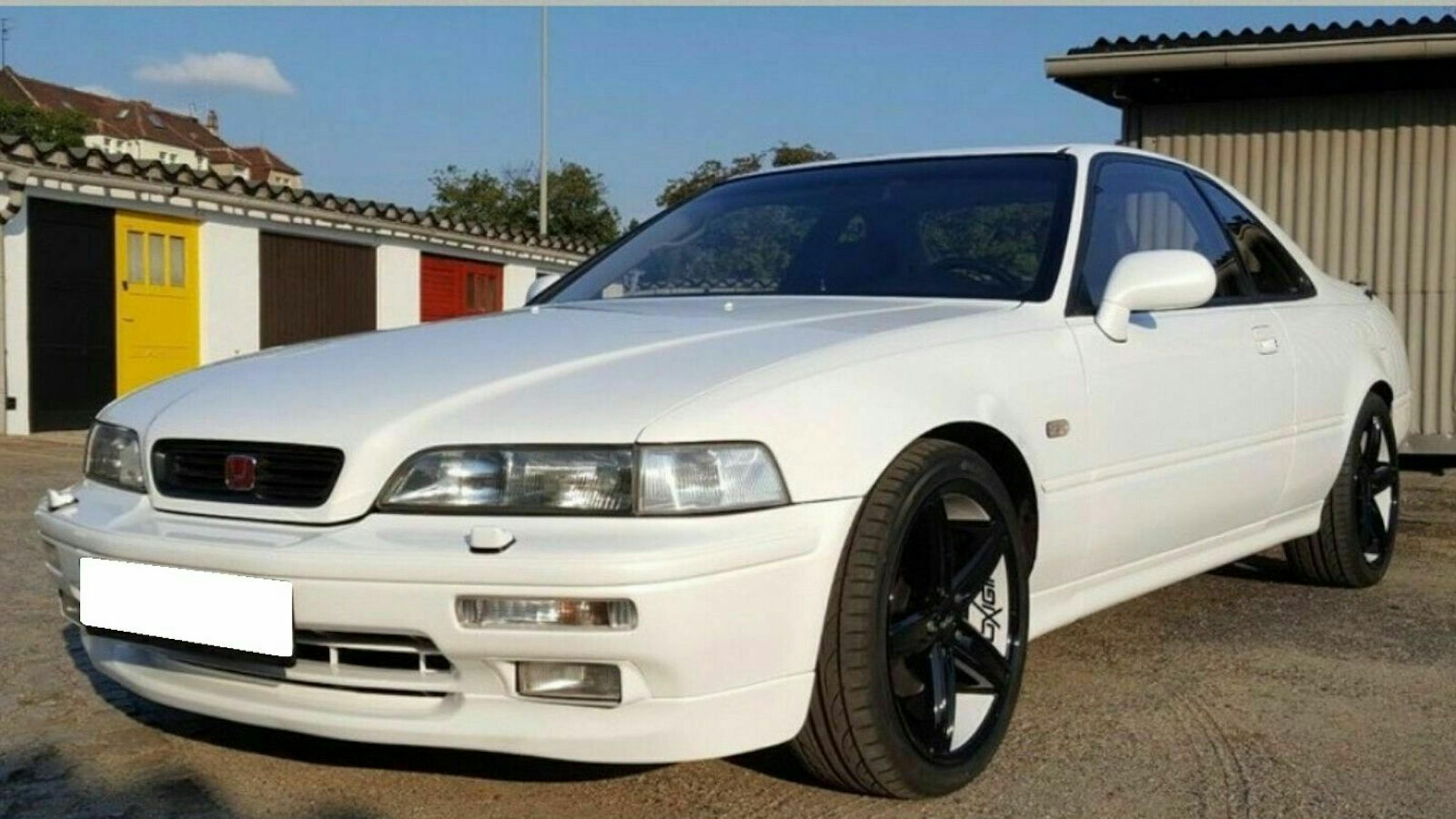 Weißes Auto mit schwarzen Felgen, parkend vor einer Garage und einem Schuppen, bei Tageslicht fotografiert.