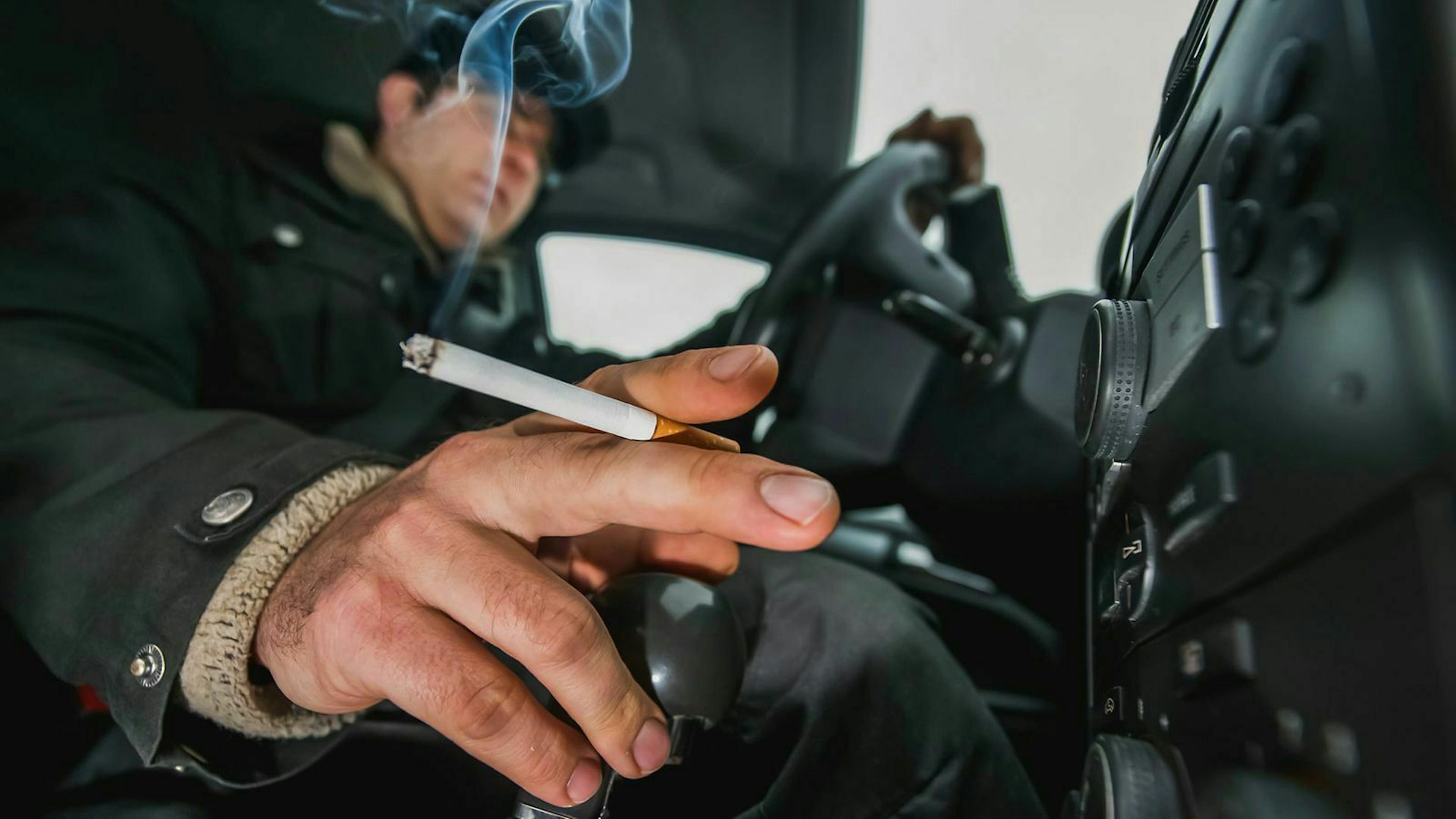 Der Fahrer eines Autos hält eine Zigarette während seine Hand auf dem Schaltknüppel ruht.