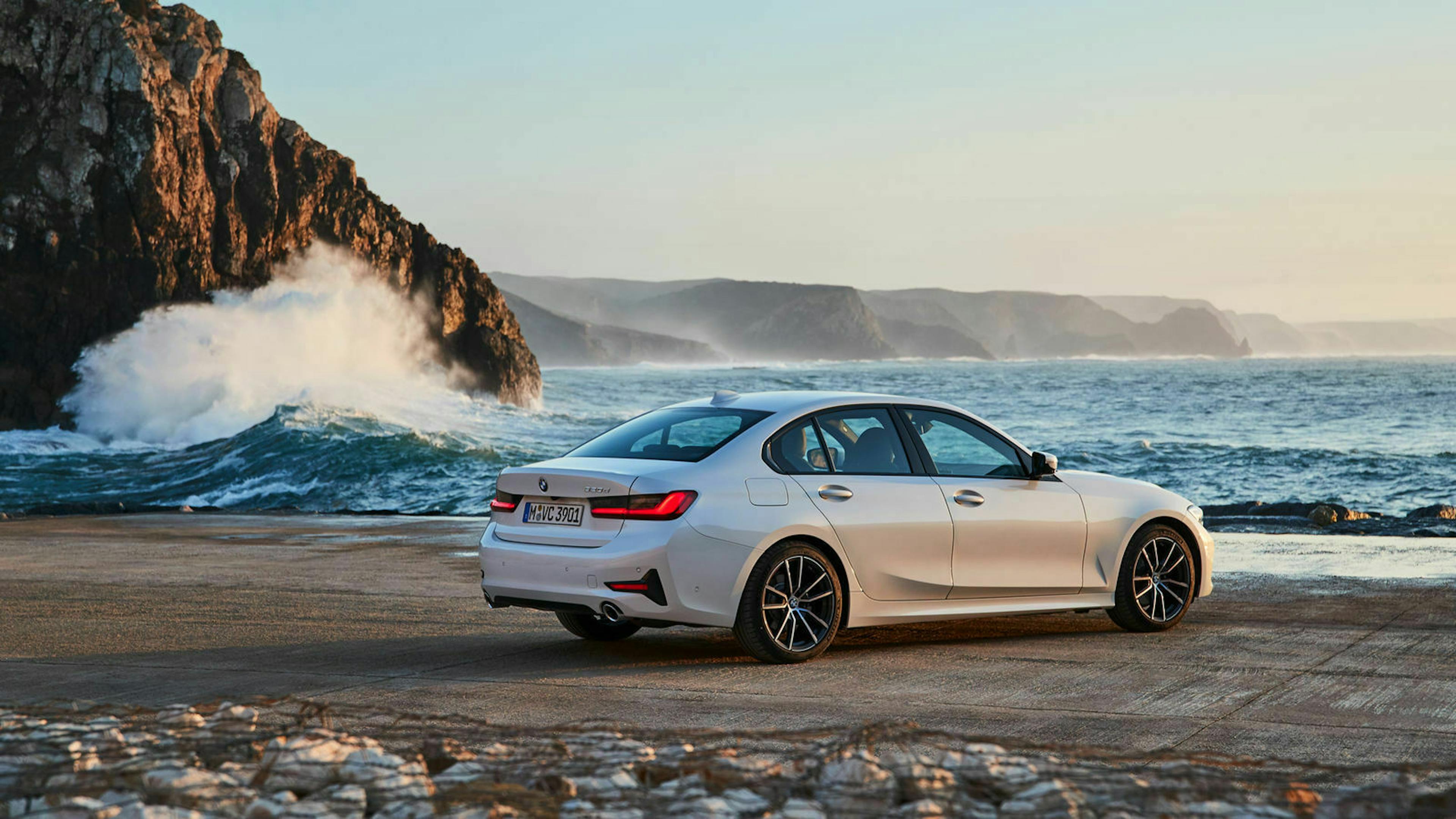 Ein weißer 3er BMW mit Heckantrieb steht auf einem Platz am Meeresufer, im Hintergrund bricht eine Welle an einer Klippe