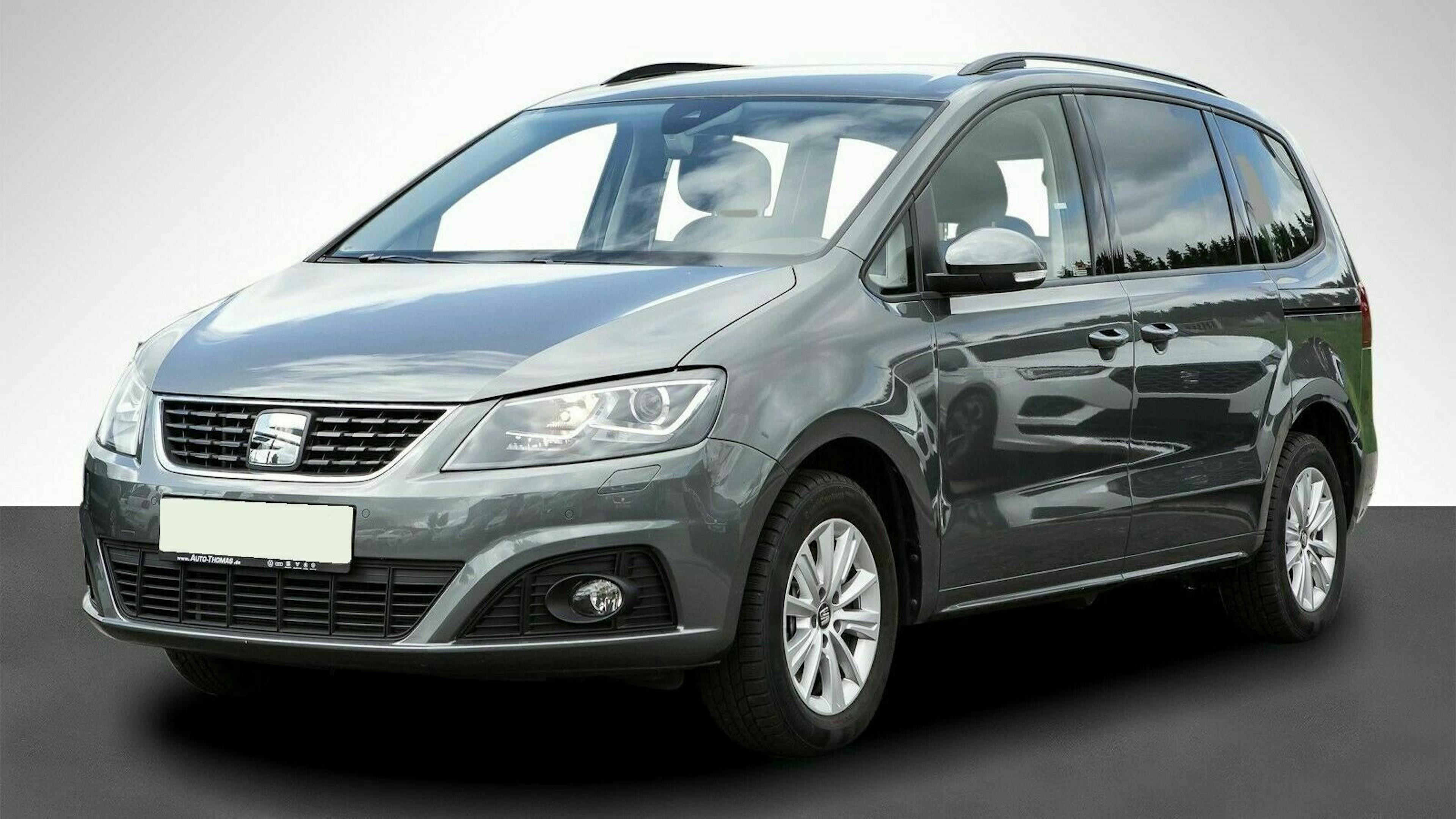 Silberner Minivan in Seitenansicht, geparkt auf dunklem Boden vor einem hellen Hintergrund. Vorder- und Seitenansicht sind sichtbar.