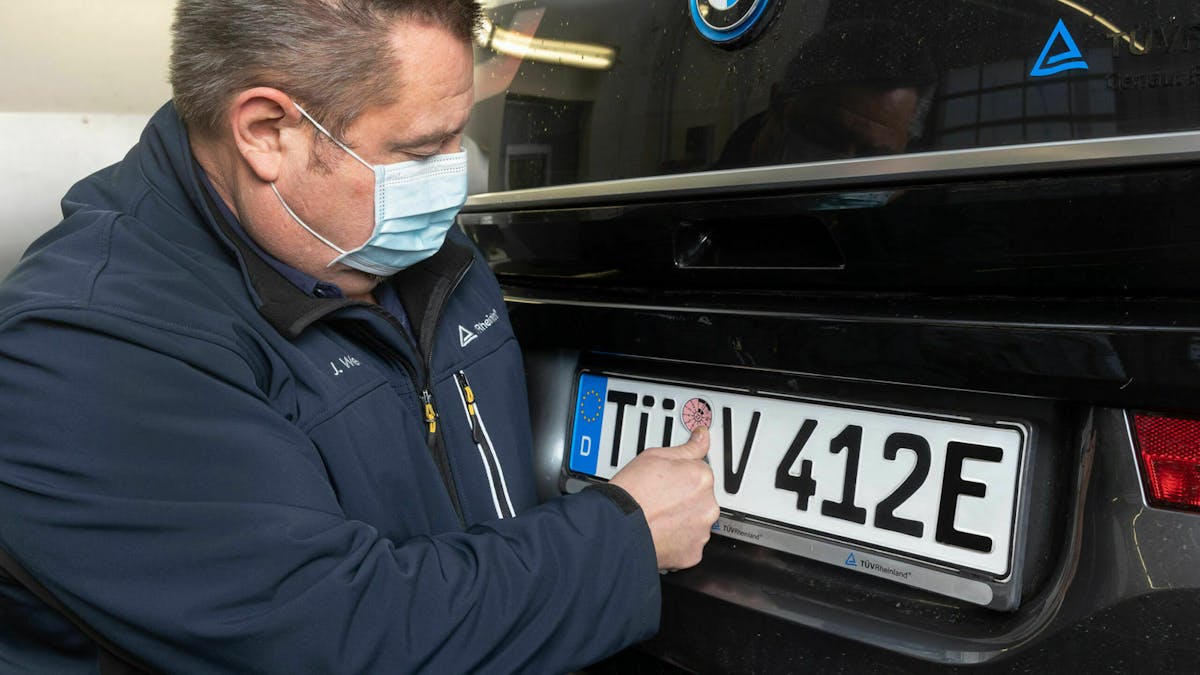 Ein TÜV-Mitarbeiter bringt eine Plakette auf dem Nummernschild eines Elektroautos an