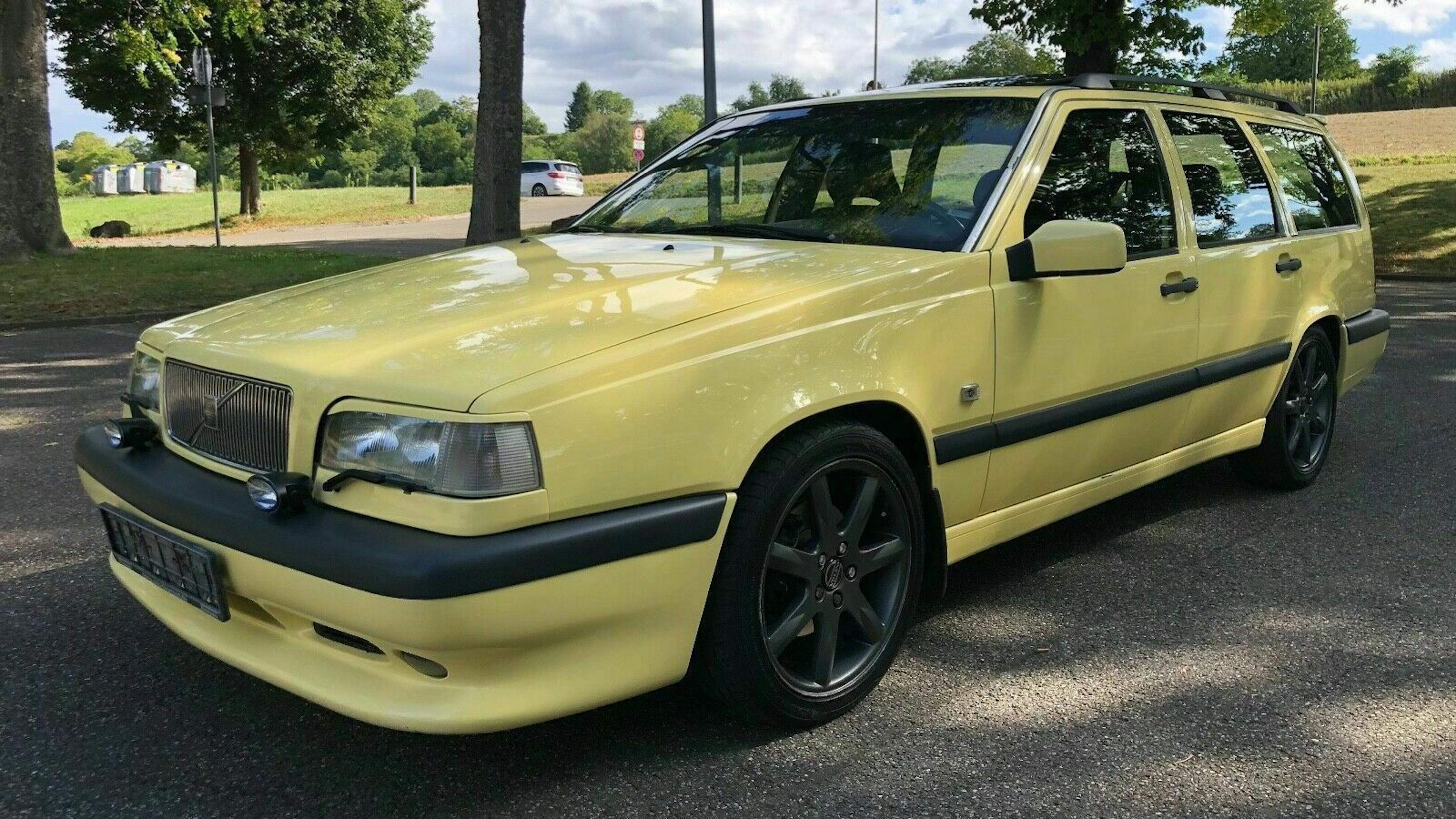 Gelber Kombi parkt auf einer Straße, umgeben von Bäumen. Der Himmel ist bewölkt. Auto hat schwarze Felgen und einen markanten Kühlergrill.
