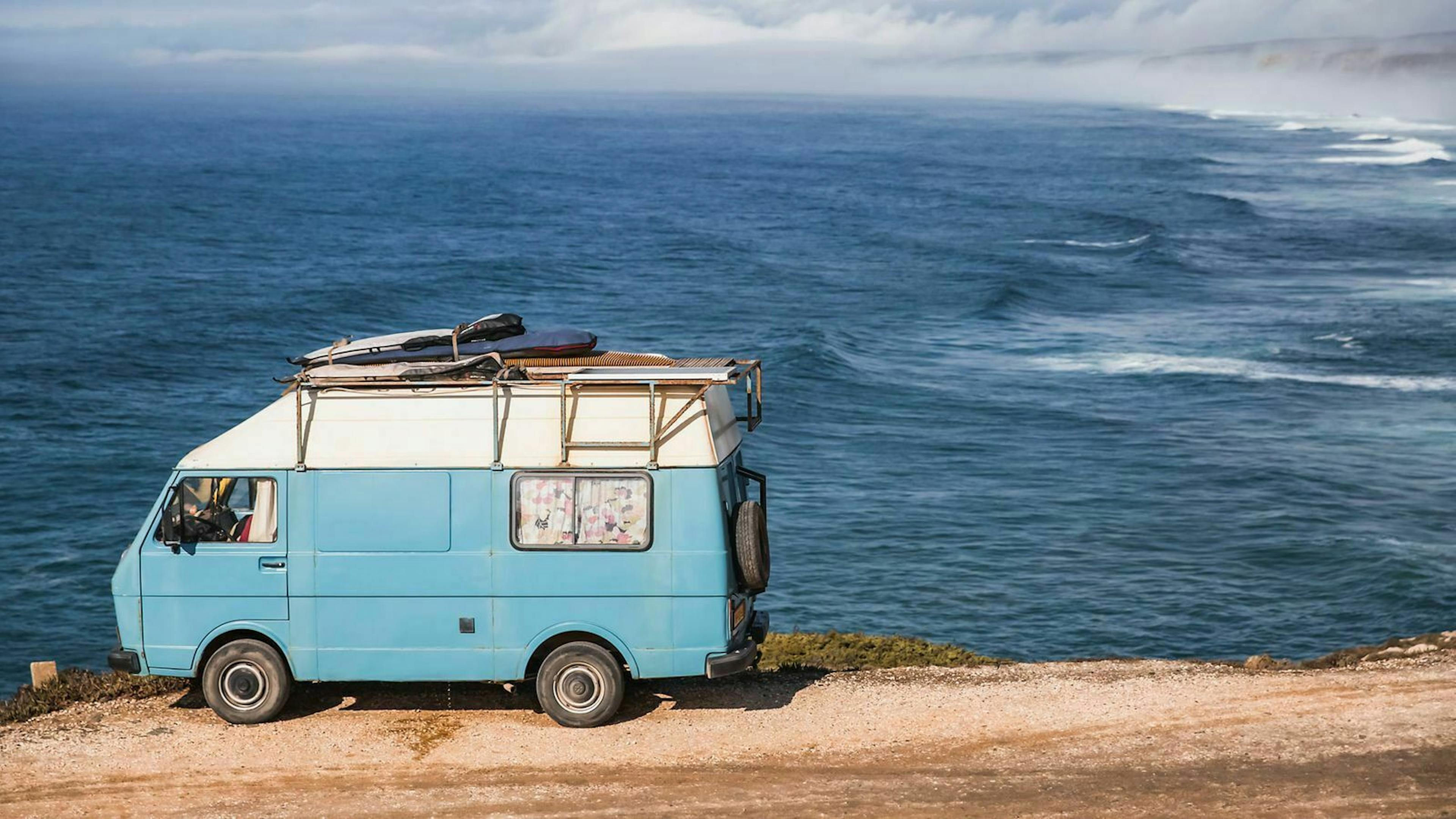 Ein hellblauer VW Camperbus steht auf einer Anhöhe über dem Meer.