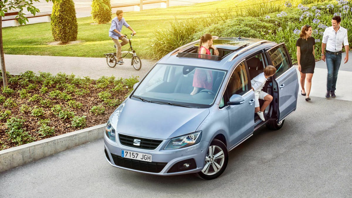 Ein blauer Seat Alhambra steht vor einem Stadtpark, zwei Kinder und zwei Erwachsene befinden sich im und vor dem Auto